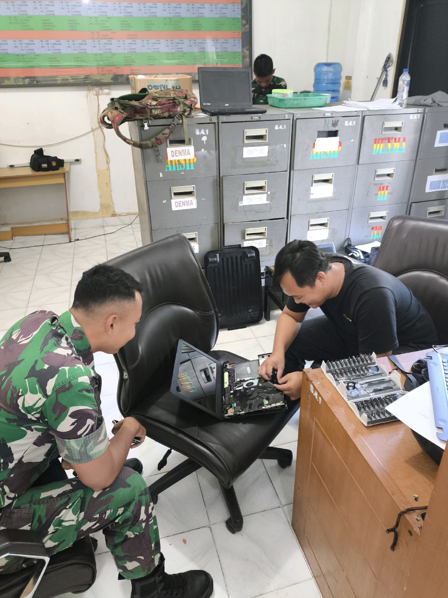 The image depicts an interior scene, likely an office or a workshop, located in an unknown city in Indonesia. Two men are present. One man, wearing a military camouflage uniform, is seated in a black leather office chair, facing a disassembled laptop which is open on another chair. He appears to be examining or working on the laptop's internal components. The second man, dressed in a black t-shirt and dark pants, is also seated and actively working on the laptop, with his hands engaged with its circuitry.
In the background, a series of grey metal filing cabinets are stacked, some with labels indicating "DENMA" and numerical designations. Above the cabinets, a large chart or bulletin board with rows of text and colored sections is visible on the wall. A laptop is open on a desk to the left, and a camouflage backpack rests on top of one of the filing cabinets. Other objects in the scene include a water cooler with a blue jug, a bottle of water, a green plastic tray, and a wooden desk with a set of precision screwdrivers and other tools laid out. The floor is tiled with white square tiles. The lighting suggests an indoor, possibly fluorescent, illumination. The overall atmosphere is one of practical work being conducted.