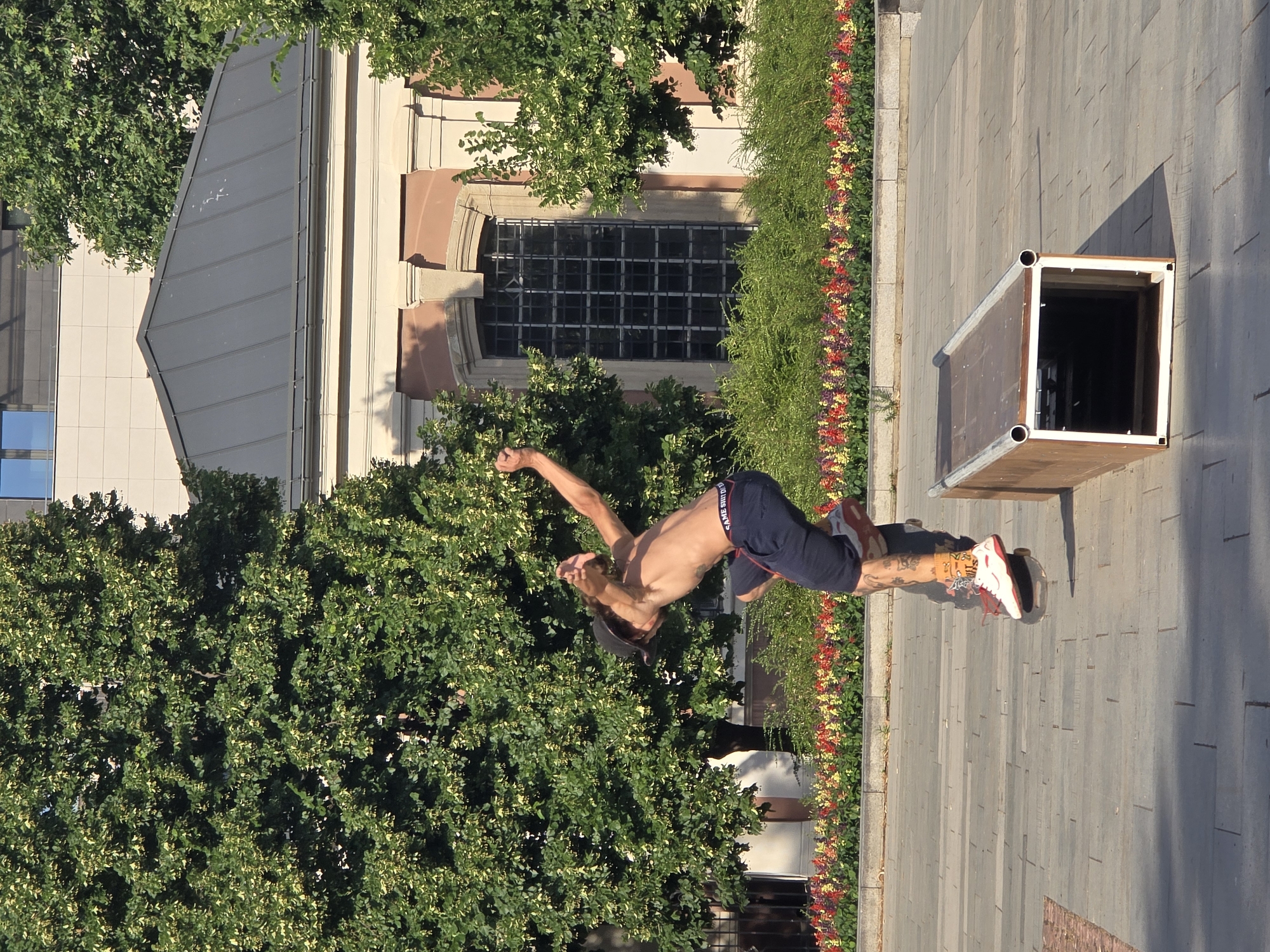 The image shows a young man performing a backflip outdoors. He is shirtless and wearing dark shorts with a red stripe and white writing. His legs are tattooed. He is wearing red and white sneakers. The man is in mid-air, with his body arched backwards. He is in front of a building with large, dark-paned windows and a grey metal roof. Lush green trees with dappled sunlight are on either side of the building. A line of colorful flowers, red, yellow, and purple, runs along the edge of a paved walkway. A large, open-topped wooden box with metal edges is also present on the walkway, casting a shadow. The scene appears to be set on a sunny day, possibly in the late afternoon due to the long shadows. The overall impression is one of athletic activity and a bright, pleasant outdoor environment. There is visible text on the man's shorts, but it is difficult to read clearly. The location is specified as Plovdiv, Bulgaria.