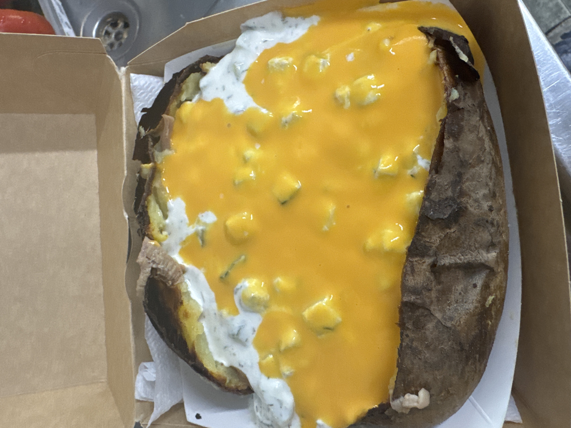 A close-up, overhead shot captures a generously topped baked potato served in a brown cardboard take-out box. The potato is split open, revealing its fluffy interior, and covered with a thick layer of bright orange-yellow melted cheese sauce. Mixed within the cheese sauce are numerous small, lighter yellow or white chunks, possibly pieces of cheese or another ingredient. A contrasting white, creamy sauce, appearing similar to sour cream or ranch dressing with visible green specks (likely herbs), is also spread over the potato, particularly on the left side, partially covered by the cheese sauce.
The potato skin is dark brown and appears crispy from baking. A white paper napkin is tucked underneath the potato within the box. The cardboard box itself is plain brown, suggesting a casual meal or take-away service. In the blurred background, a metallic kitchen sink with a circular drain is visible, along with a red, rounded object, possibly a tomato. The setting appears to be indoors, likely a kitchen counter. No specific activity is depicted, but the prepared food suggests it is ready for consumption. There is no visible text in the image. The photo was taken in Нареченски бани, Bulgaria.