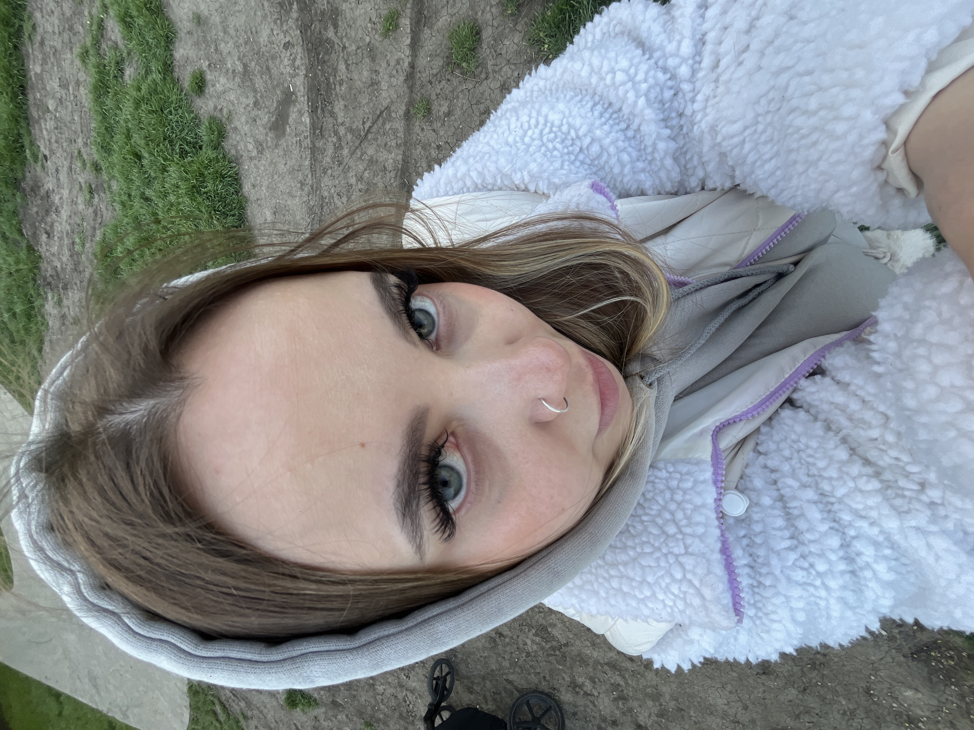 A selfie captures a young woman from an overhead angle, with the image rotated 90 degrees clockwise. She has light brown hair with blonde highlights, clear skin, prominent eyelashes, and striking green or blue eyes that look directly at the viewer. A silver hoop nose ring is visible on her right nostril.
She is dressed in warm attire, featuring a white or cream-colored fluffy sherpa jacket with a textured surface. Underneath, she wears a gray hooded sweatshirt, which has a light purple zipper detail running down the front. The hood of the sweatshirt frames her face.
The background depicts an outdoor setting, characterized by patches of vibrant green grass interspersed with areas of dry, cracked earth or dirt pathways. This ground surface is visible beneath her, suggesting she is outdoors, possibly looking down or lying on the ground while taking the photo. A portion of what appears to be a black wheel, possibly from a stroller or similar wheeled object, is visible at the very bottom left corner of the frame.
The natural lighting indicates it is daytime. The combination of a warm jacket and the outdoor environment, featuring grass and earth, suggests she is in an informal outdoor area, possibly a park or walking path in Krasnodar, Russia. No visible text is present in the image.