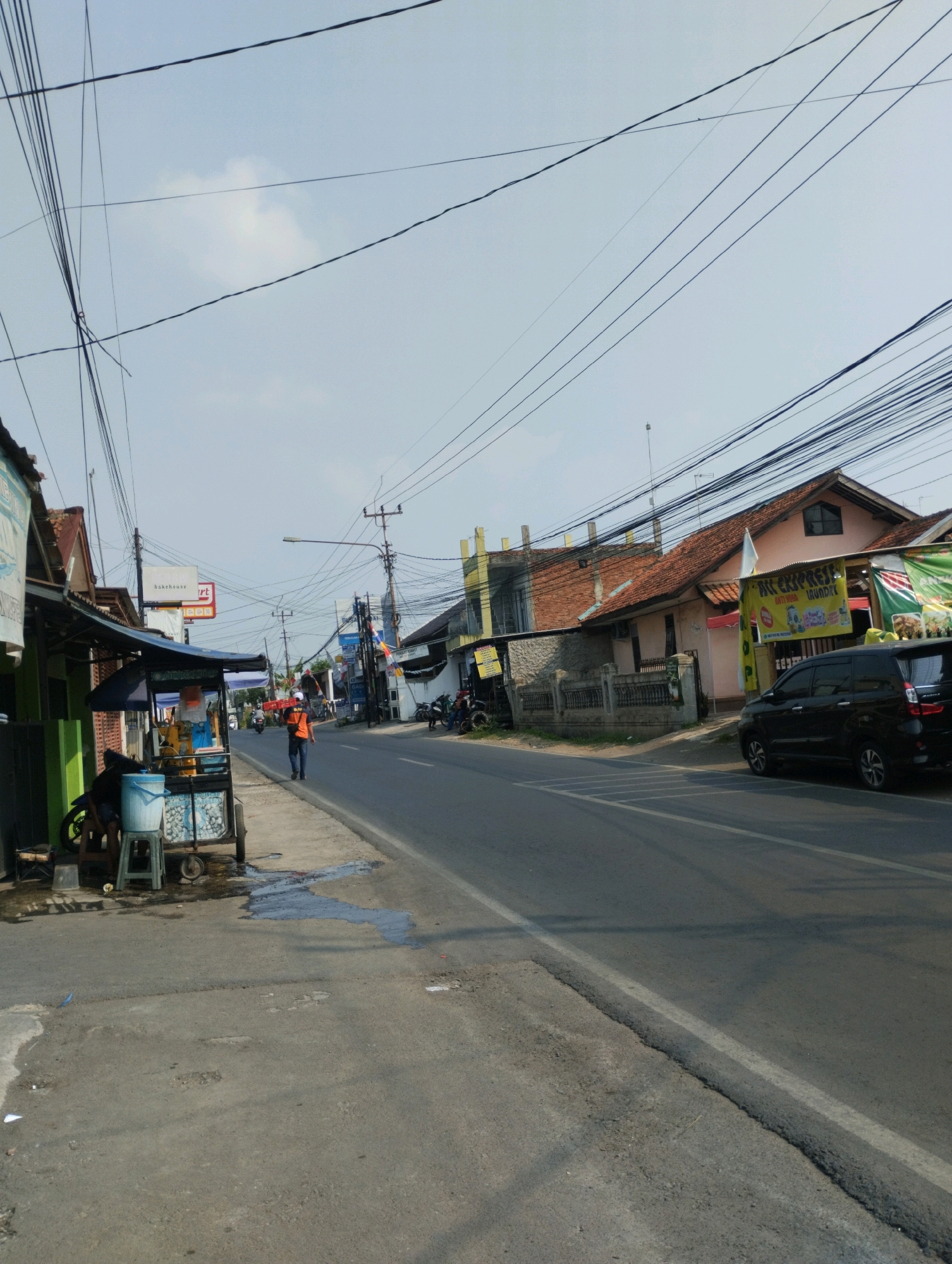 This image depicts a street scene in Purwakarta, Indonesia, characterized by a clear sky and overhead power lines. The road is paved and has a white line marking the lane. On the left side of the road, there's a sidewalk with a food cart and a parked motorcycle. A person wearing an orange vest is walking on the sidewalk. Adjacent to the sidewalk are several buildings, including a business with a sign that reads "bakehouse art." Further down the street, more buildings line the road, with another business displaying a yellow banner with Indonesian text. A black SUV is parked on the right side of the street in front of a building with a red-tiled roof. The environment appears to be a mix of residential and commercial properties.