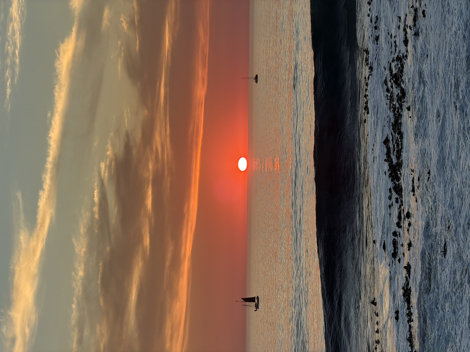 A dramatic sunset over the ocean casts a vibrant orange glow across the sky and sea, with the sun a bright red orb sinking towards the horizon. In the foreground, the ocean churns with white foam and dark seaweed-covered rocks. Two sailboats are visible in the distance. The image appears to be taken from a beach or rocky outcrop overlooking the sea.
While the image depicts a sunset over the ocean with sailboats, it is impossible to definitively determine the specific location in Cape Town, South Africa, or to provide further details about the scene without additional information or context.