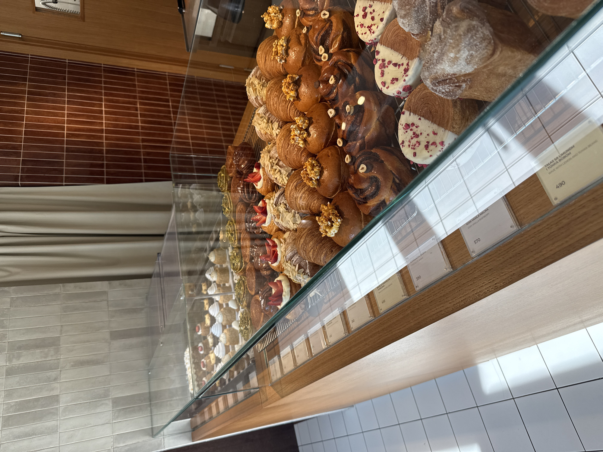 An overhead, slightly angled view captures an illuminated glass display case filled with a diverse array of pastries and baked goods, likely in a bakery or patisserie in Belgrade, Serbia.
The central focus is on numerous croissant-like pastries artfully stacked. These include variations topped with whole hazelnuts, crushed nuts, or caramel popcorn-like pieces. Other pastries feature a half-dip in white icing adorned with red sprinkles, suggestive of dried raspberries. Further into the display, more intricate desserts are visible, such as pastries with fresh strawberry slices, and smaller, individual cakes or tarts topped with green pistachios or white frosting.
Small, rectangular price tags are arranged along the base of the display case, featuring descriptions in both Serbian and English. Visible text includes:
* "TALAS SA LIMUNOM I TONKA BOBOM" (Wave with Lemon and Tonka Bean) priced at 490.
* "KOIN VANILA MALINA" (Coin Vanilla Raspberry) priced at 570.
* "CHOCOLATE BABKA" priced at 390.
* A partially visible label mentions "RASPBERRY" and "POPCORN CARAMEL," priced at 450. Another tag shows a price of 260. The prices suggest Serbian Dinars.
The background features dark brown horizontal subway tiles on the left, a light wood panel above the display, and white square floor tiles on the right. The bright, even lighting suggests an indoor commercial setting during the daytime. No people are visible, but the meticulously arranged display indicates a retail environment prepared for customers.