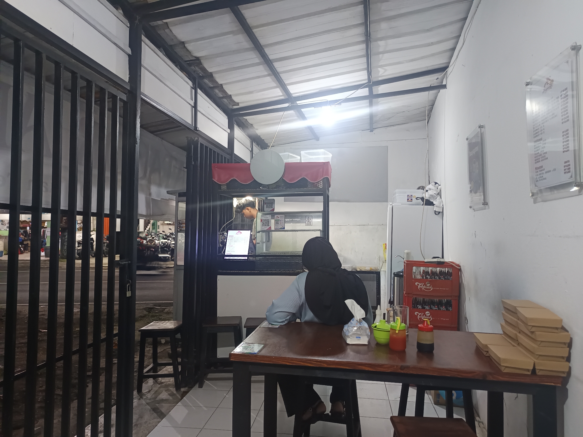 The image depicts the interior of a casual dining establishment in Bogor, Indonesia, likely a warung makan or small restaurant. A metal gate with vertical bars is visible in the foreground on the left, and a metal roof structure supported by beams covers the space. The lighting is artificial, with a prominent bulb overhead.
Inside, a person wearing a black hijab and a light blue shirt is seated at a dark wooden table, facing away from the camera. They are looking towards a counter where a young man in a green shirt is visible, possibly preparing food or taking an order. The counter area has a shelf with a red valance, a circular object, and several clear plastic containers. A laptop is also on the counter, displaying text.
On the table in front of the seated person are several items, including a tissue box, two squeeze bottles (one red and one brown), and a green dispenser. To the right of the table, a stack of brown cardboard food boxes is neatly piled. Behind the counter, a white refrigerator and red crates with the "Teh Botol" logo are visible.
The background, seen through the open gate, shows a street scene with parked motorcycles and vehicles, suggesting the establishment is located on or near a road. The overall impression is of a simple, functional eatery.