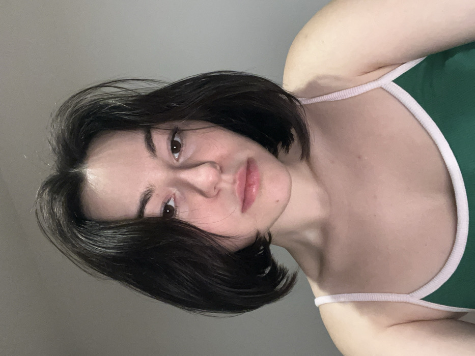 A young woman is depicted from a low angle. She has dark, short, slightly wavy hair and fair skin. She wears a green tank top with white straps. Her expression is serene, with full lips and dark eyes. The background is a muted gray, putting the focus entirely on the woman. The lighting is soft, creating subtle shadows on her face and neck.