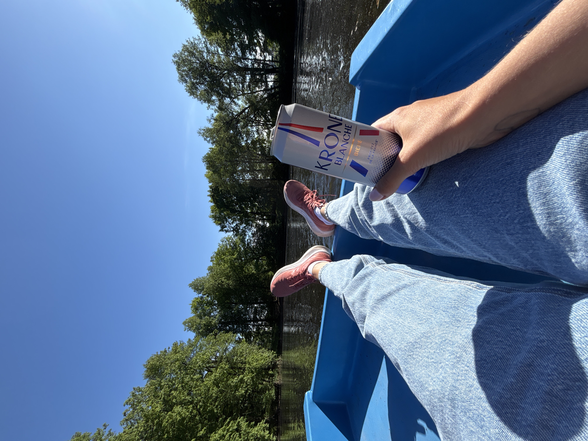An outdoor, first-person perspective shot captures a person relaxing in a bright blue boat on a sunny day. The person's lower body is visible, dressed in light blue denim jeans and wearing pink athletic shoes. Their right hand holds a silver can of "Kronenbourg Blanche" beer, with the word "BIÈRE" and the phrase "PROFITĒZ DE LA FRANCE" clearly legible on the label.
The boat floats on a calm body of water, likely a river or canal, reflecting the surrounding greenery. Dense, leafy trees line the banks in the background, suggesting a natural or park-like environment. Above, the sky is a vast expanse of clear, vibrant blue with no clouds, indicating excellent weather.
The bright sunlight illuminates the scene, casting soft shadows inside the boat and on the person's jeans. The overall mood conveyed is one of leisure and enjoyment, perhaps during a vacation or a relaxing weekend outing. The angle emphasizes the tranquil setting and the simple pleasure of a drink on the water.