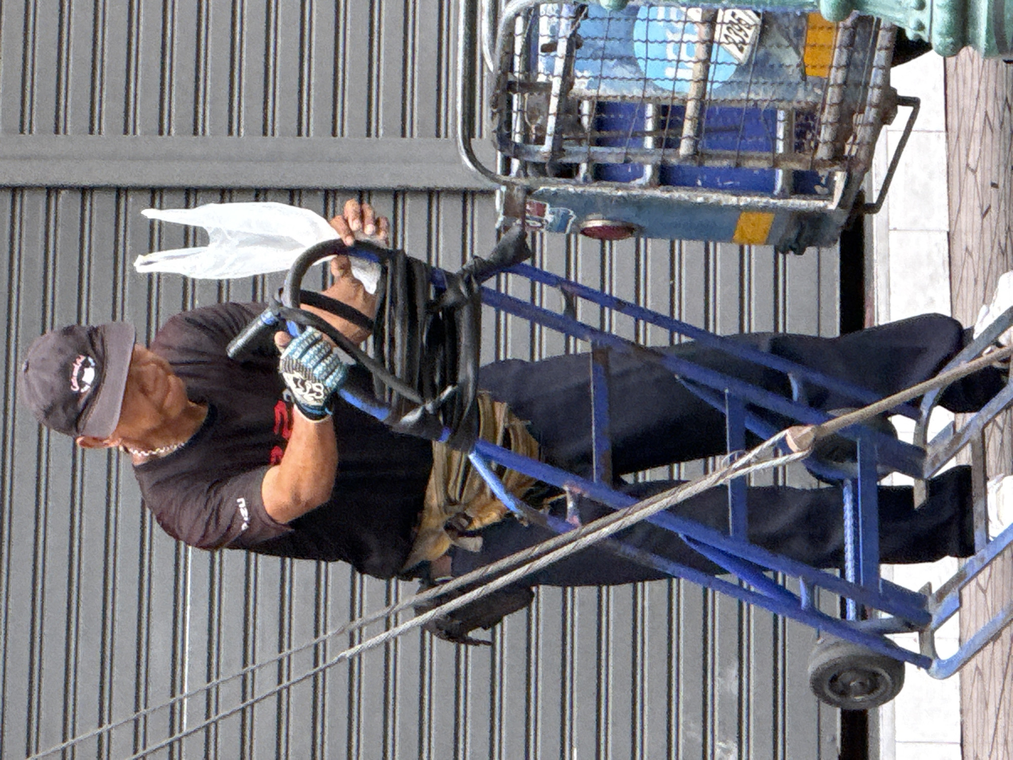 A man wearing a black t-shirt, dark pants, a black baseball cap, and protective gloves is standing on a sidewalk in Bangkok, Thailand. He is holding a blue hand truck with a black bag attached to it. A white plastic bag is held in his right hand. The background is a grey corrugated metal door.