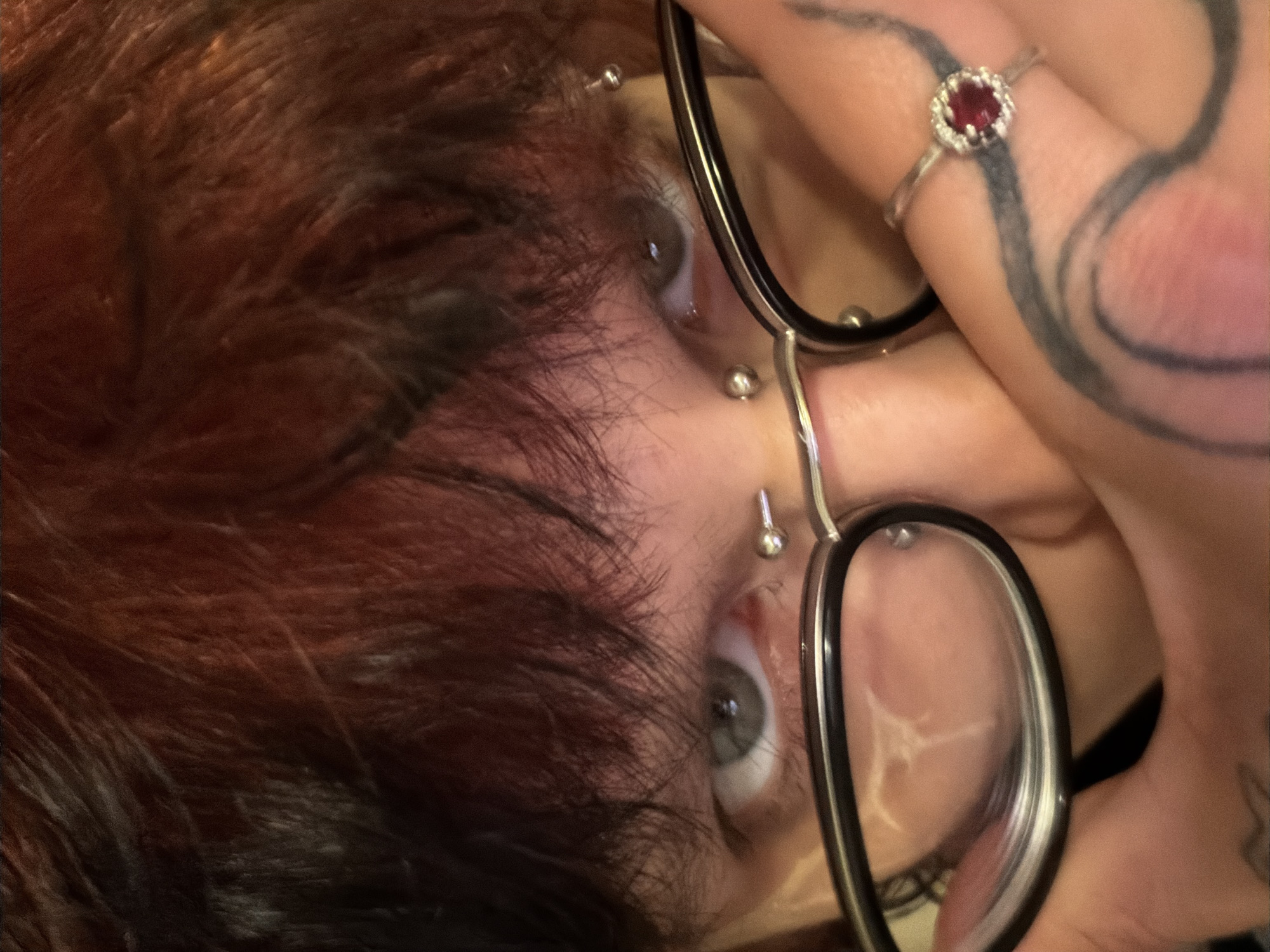 An eye-level close-up, rotated, shows a person holding glasses. The person has red hair with dark roots and fair skin, with two piercings on their face and another between their eyes. They are wearing a ring with a red gemstone. The fingers holding the glasses have a tattoo of a spiral. A scar is visible on the face behind the lenses of the glasses. The location of this image is unknown and cannot be determined from the visual data provided.