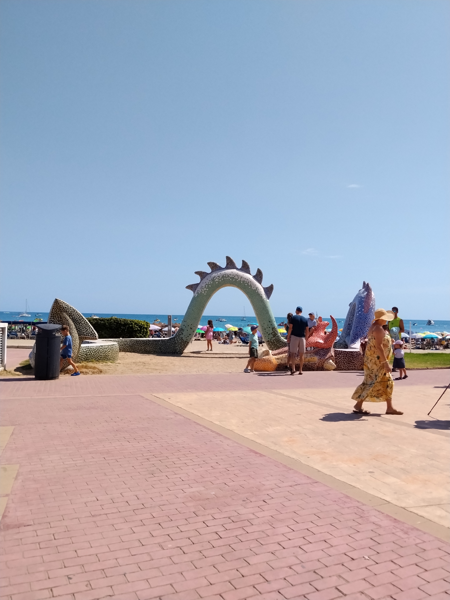 A wide, low-angle shot captures a sunny day at the beach in Peníscola, Spain. In the foreground, a red brick walkway leads the eye towards the beach and a mosaic sculpture of a dragon. The dragon sculpture is a vibrant green with a spiky back, forming an archway over a sandy area. To the right of the dragon, a blue mosaic dolphin-like sculpture is visible. Several people are scattered around the dragon sculpture, some standing, some sitting, and children are playing near the beach.
Beyond the sculpture, the beach is bustling with activity. Numerous beach umbrellas are set up on the sand, and people are relaxing and walking. The blue ocean stretches to the horizon, dotted with sailboats. The sky is a clear, bright blue, indicating fair weather. On the left side of the image, a black trash bin stands near the walkway. A young boy in blue shorts and a t-shirt is walking past the trash bin. To the right of the dragon sculpture, a woman in a yellow floral dress and a straw hat walks away from the camera.
The overall scene depicts a lively and colorful beachside resort.