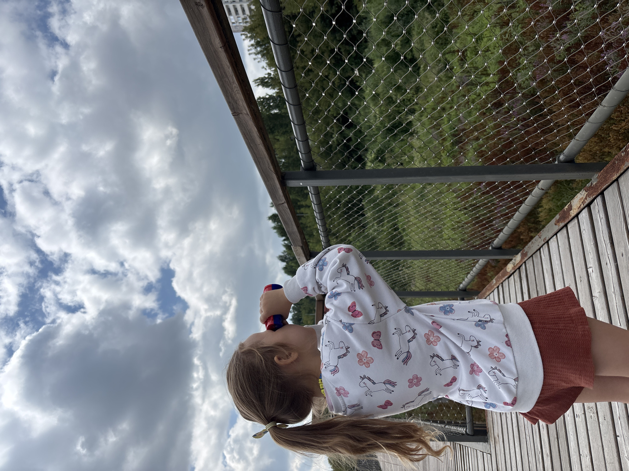 A young girl with blonde hair in a ponytail, wearing a white sweatshirt with unicorn and flower patterns and rust-colored shorts, stands on a wooden walkway and looks through red and blue binoculars at a cloudy sky. She is positioned on the right side of the frame, facing away from the viewer and towards the right. A metal mesh fence with a wooden railing runs along the right side of the walkway, separating it from a natural area with green trees and some vegetation. A tall residential building is visible in the background on the far right. The wooden walkway is made of planks laid horizontally. The overall scene suggests an outdoor setting, possibly a park or elevated boardwalk, within the city of Orenburg, Russia.