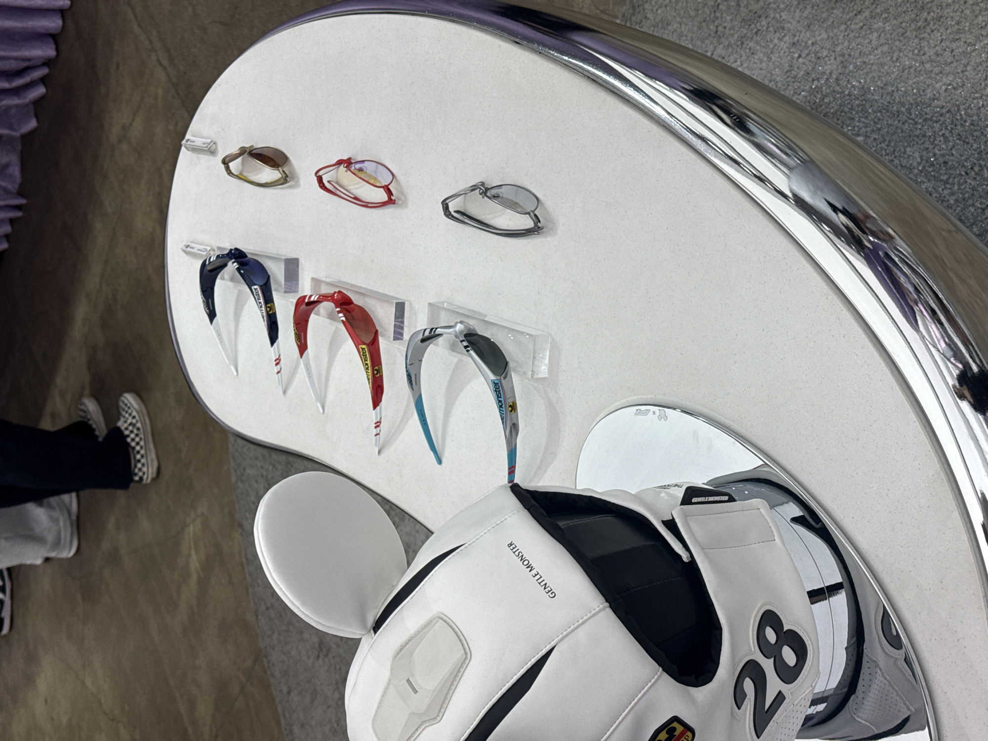 The image shows a display case of Gentle Monster sunglasses, likely in a store location in Seoul, South Korea. The display is white with a curved edge and a mirrored accent. Three pairs of sunglasses are resting directly on the surface, while three others are mounted on clear plastic stands. Below the display, partially visible, is a white helmet with black accents and the number "28" printed on the side, also appearing to be a Gentle Monster product. To the left, the lower legs and feet of a person wearing dark pants and checkered Vans sneakers are visible. The floor appears to be a mix of light brown and gray tones.