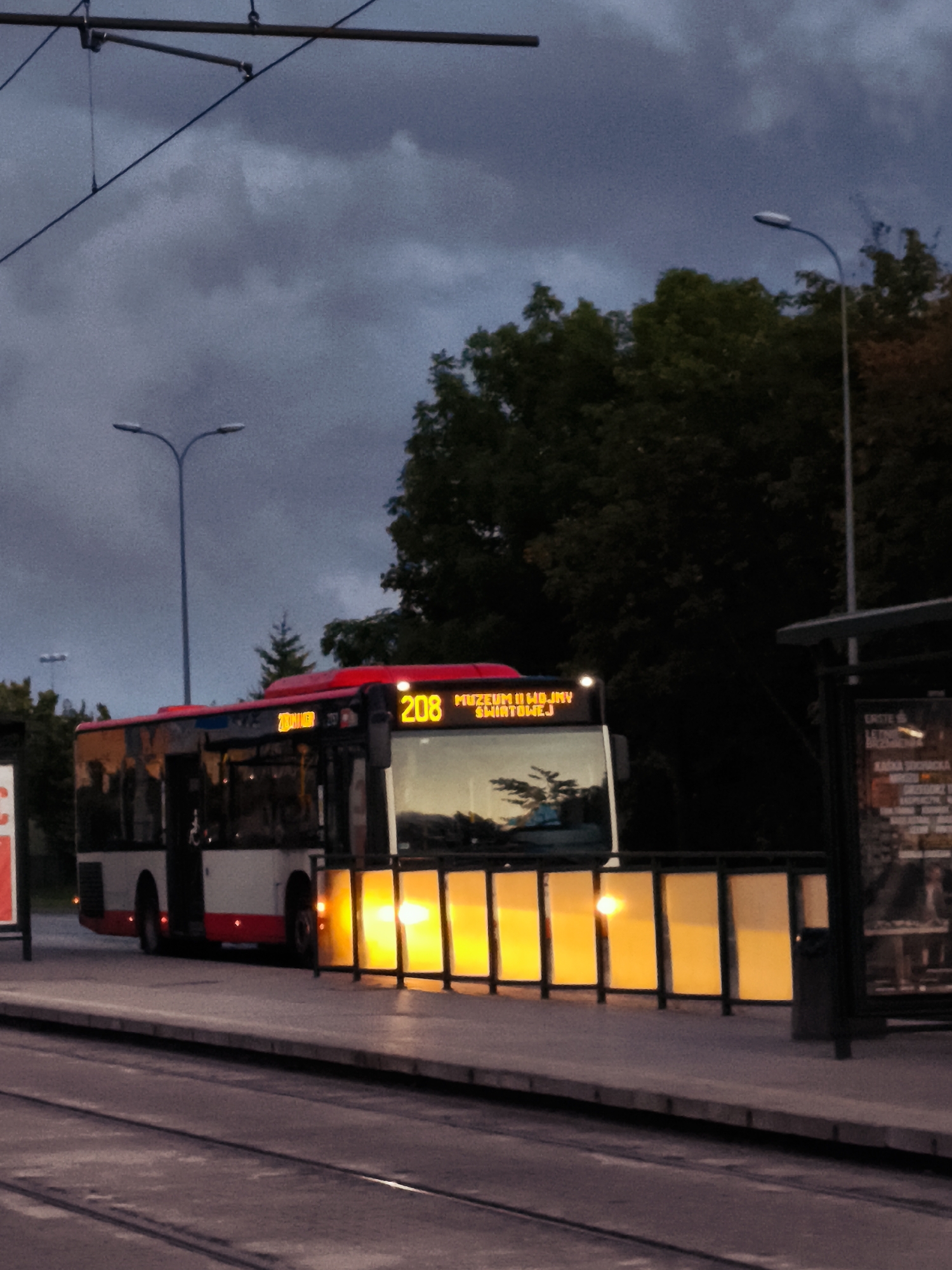 The image captures a public transportation scene in Gdańsk, Poland, likely at a tram or bus stop. A red and white bus, displaying the route number "208" in illuminated orange digits, is positioned at the stop. The bus has its headlights on, and its destination is visible as "MUZEUM II WOJ. SWIATOWEJ". This translates to "Museum of the Second World War," a prominent landmark in Gdańsk, confirming the location. The bus has its doors open, indicating it is either boarding or alighting passengers, although no individuals are clearly visible in the process.
The environment is characterized by a cloudy, twilight sky, suggesting it is either early morning or late evening. The bus stop features a modern shelter with illuminated panels emitting a warm yellow light, contrasting with the dim ambient light. Tram tracks are visible in the foreground, running parallel to the bus and the platform. Streetlights, one with a distinctive double-headed design, are present in the background, along with trees and overhead power lines for the trams. A poster board or advertisement is affixed to a post near the bus shelter. The overall atmosphere is calm, with the focus on the bus and its operational status at the stop.