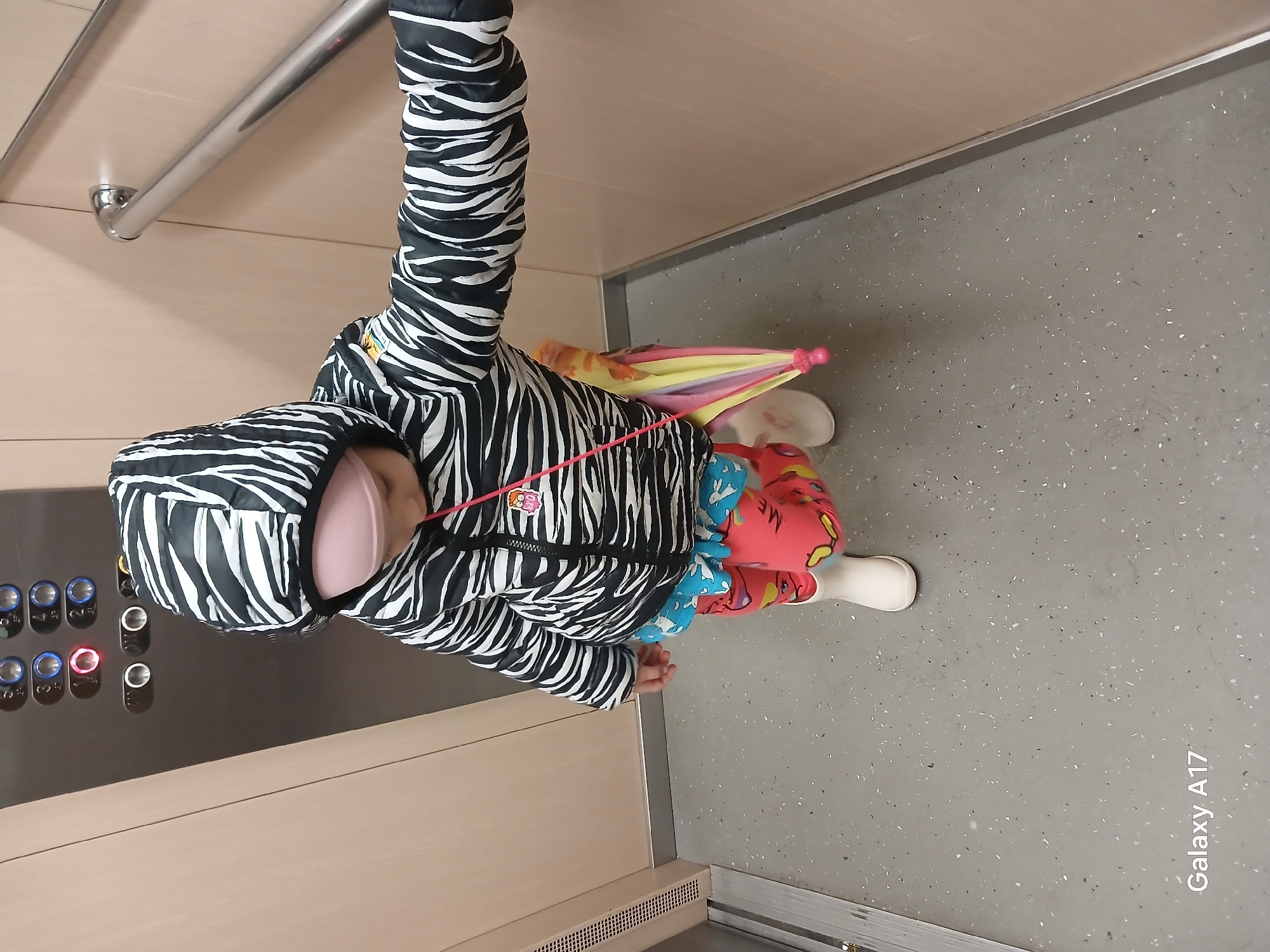 The image shows a child standing in what appears to be an elevator. The child is wearing a black and white zebra-striped jacket with a hood, and colorful leggings with a cartoon-like pattern. They are also wearing white boots. Next to the child is a small, closed, multicolored umbrella. To the left, there is a control panel with numerous buttons. A handrail is visible in the upper left corner. The elevator's interior consists of light beige wall panels and a gray floor. "Galaxy A17" is visible as a watermark at the bottom right. The image was most likely taken in an elevator. There is no evidence indicating the location is Yekaterinburg, Russia.