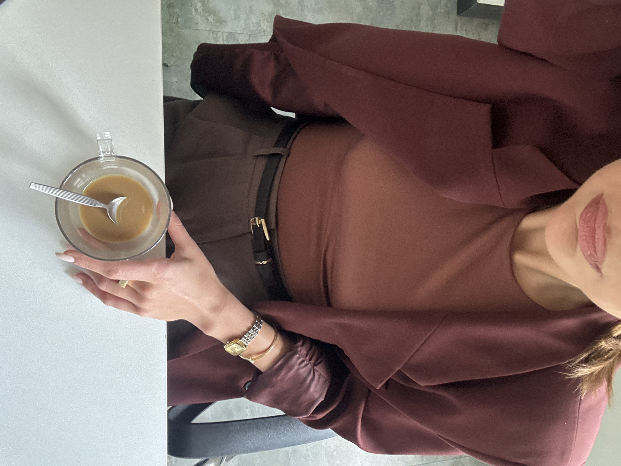 A woman is seated at a white table, holding a glass mug of coffee. She wears a brown top, brown pants with a black belt and gold buckle, and a maroon blazer. Jewelry includes a gold ring, a watch, and a gold bracelet. The location is unclear.