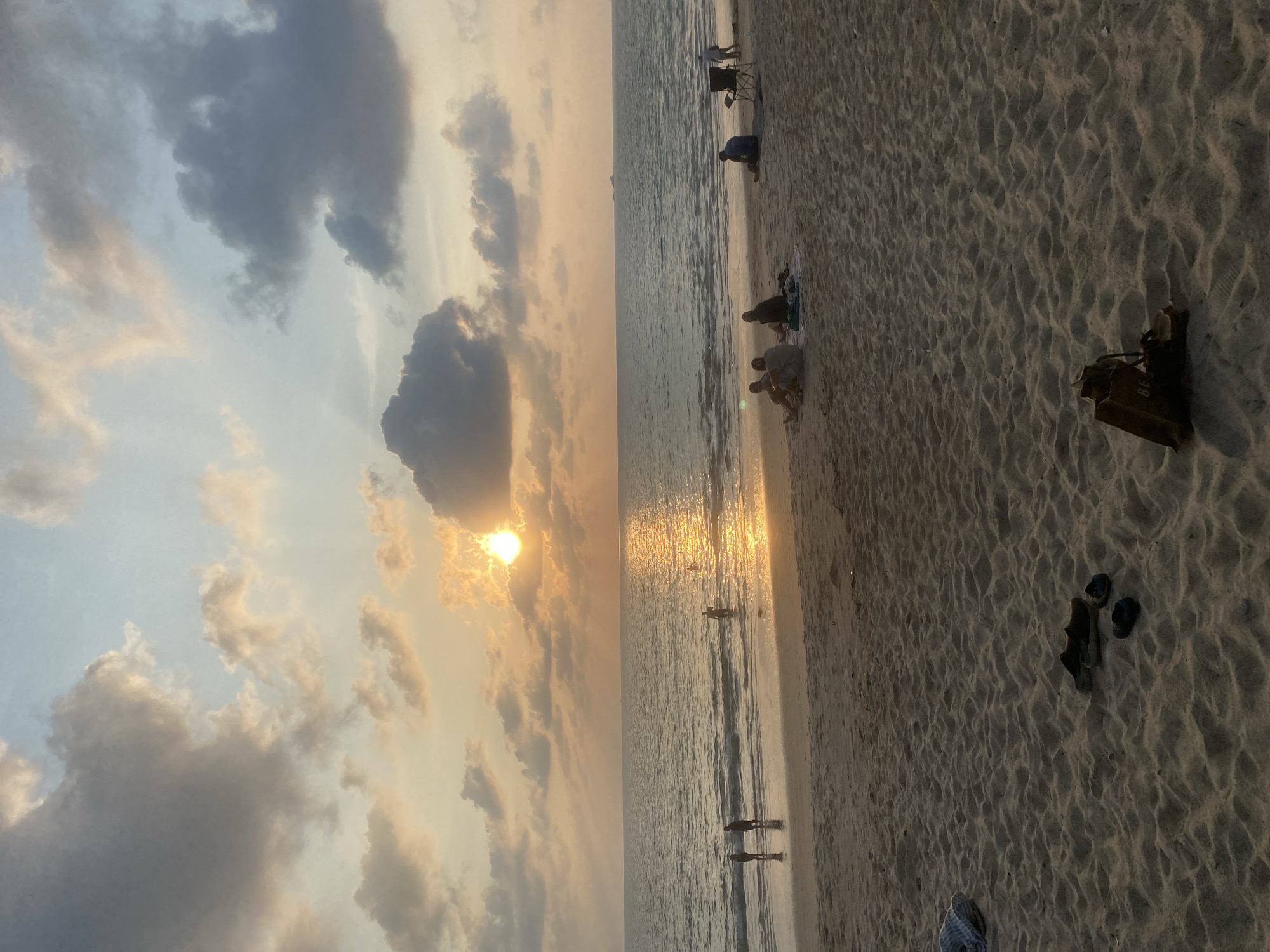 The image displays a serene beach scene at sunset in Choeng Thale, Thailand. The sun, a bright golden orb, is low on the horizon and partially obscured by scattered clouds, casting a warm, radiant glow across the sky and reflecting intensely on the surface of the sea. The sky presents a blend of soft blues, grays, and warm oranges and yellows, with various clouds catching the last light of the day.
Along the sandy shore, several people are present. Some individuals are seen sitting or crouching near the waterline, while others are standing or wading in the shallow part of the sea, their figures silhouetted against the bright reflection on the water. The beach itself is characterized by rippled sand textures. Various objects are visible on the sand, including an empty beach chair, a dark bag or backpack with some faint text (possibly "B.E.L." or "B.F.L."), and a pair of dark flip-flops or sandals.
The time of day is clearly sunset, indicated by the position of the sun and the warm, soft lighting. The weather appears calm with partly cloudy skies. The overall atmosphere suggests a peaceful and relaxing evening by the sea.