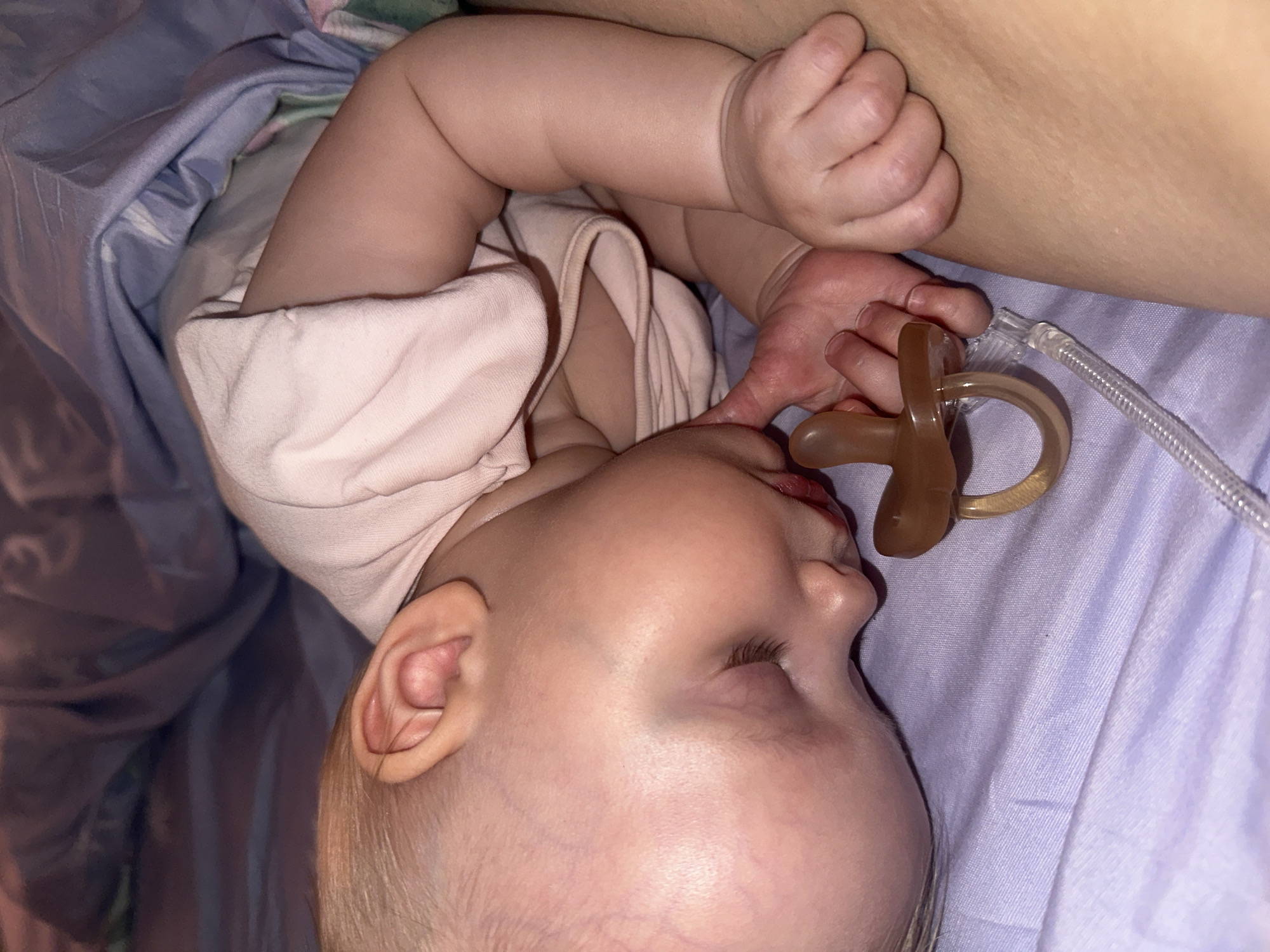 A baby is lying on their side in a bed. The baby has fair skin, blonde hair, and closed eyes. They have a pacifier in their mouth and one hand is touching it. The baby is wearing a light pink shirt. The bed has a light purple sheet and a blue blanket. There is no indication that this image was taken in Pokrov, Russia.