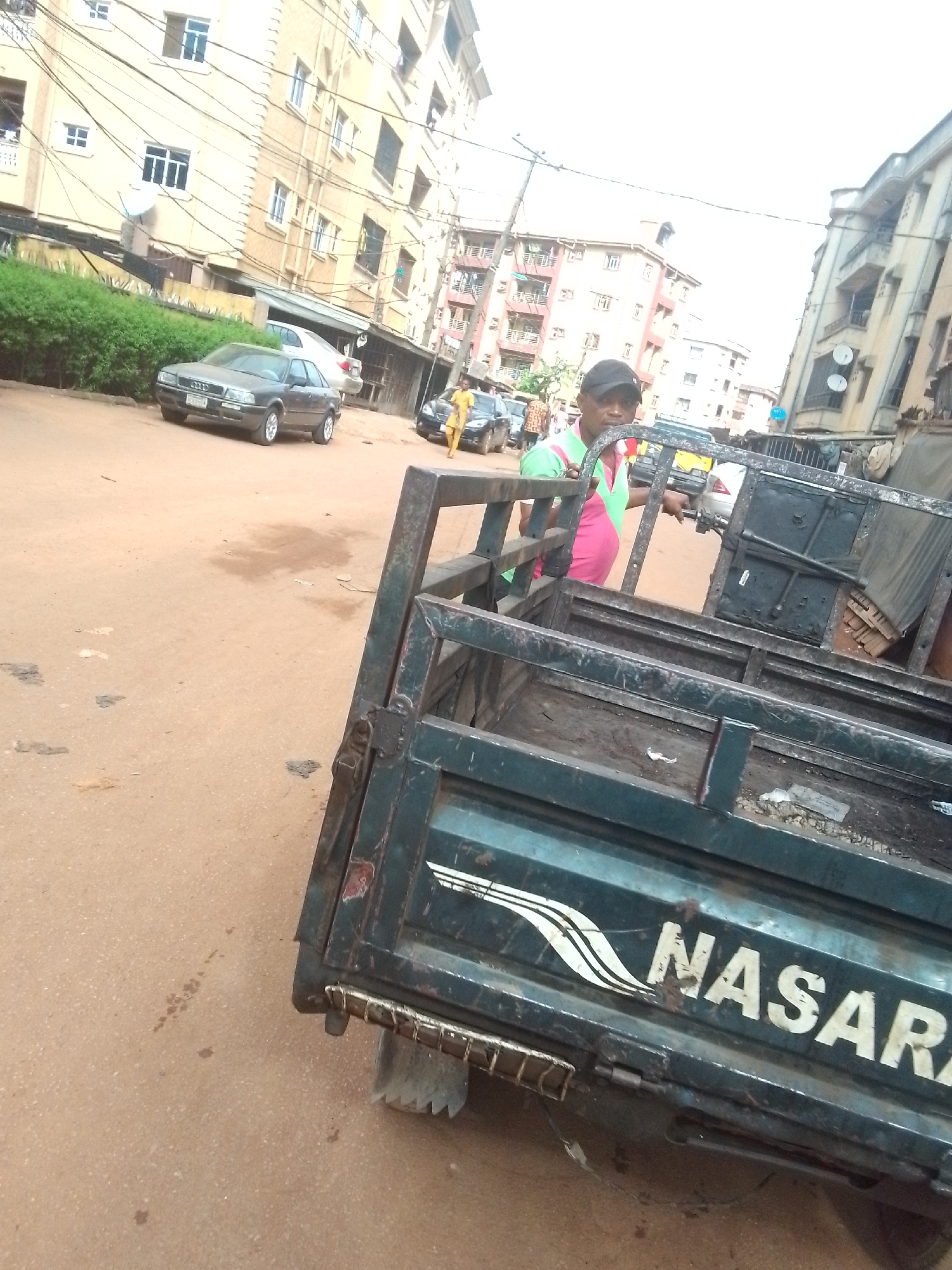 The photo shows a street in Onitsha, Nigeria. In the foreground is the back of a small truck with the name "NASARA" painted on it. A man wearing a baseball cap, green shirt, and pink vest stands behind the truck bed, facing the camera. In the background, there are several buildings and other cars parked along the street. Several people are visible walking on the sidewalks and the street. The road appears to be unpaved.
