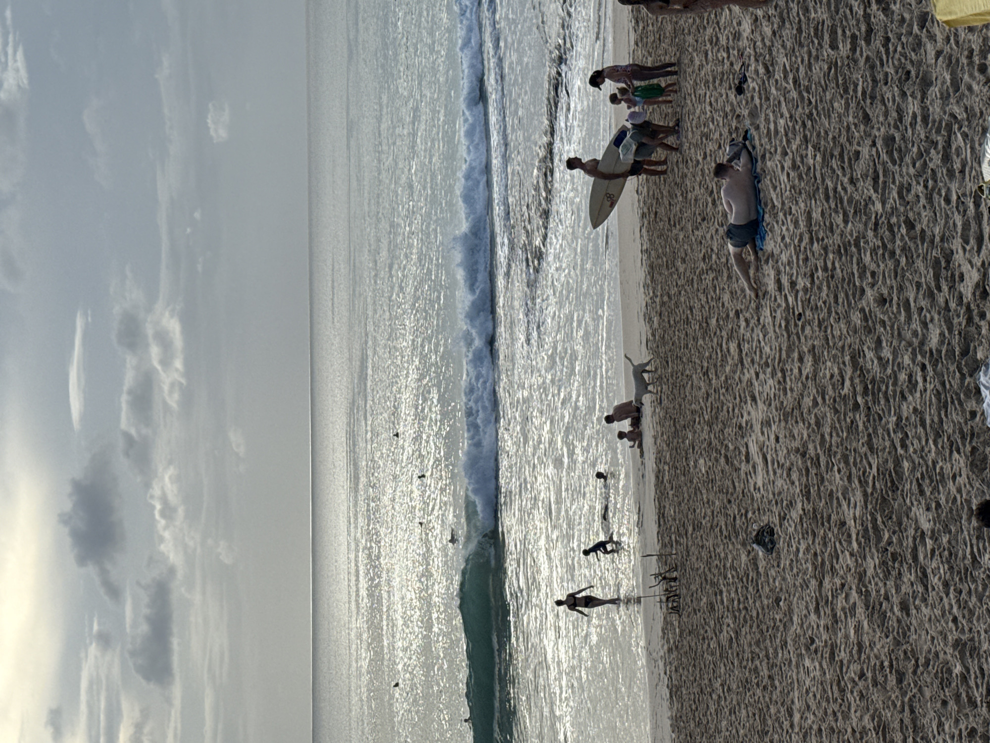 The image depicts a beach scene in Kuta Selatan, Indonesia, likely at sunset or sunrise, as evidenced by the diffused lighting and clouds in the sky. The scene is set at a beach where people are enjoying various activities. There are people swimming and people walking near the water. A group of people are together, one person holding a surfboard. One person is lying face down on a blue mat on the sand. The environment includes a large body of water with waves, a sandy beach, and a cloudy sky.