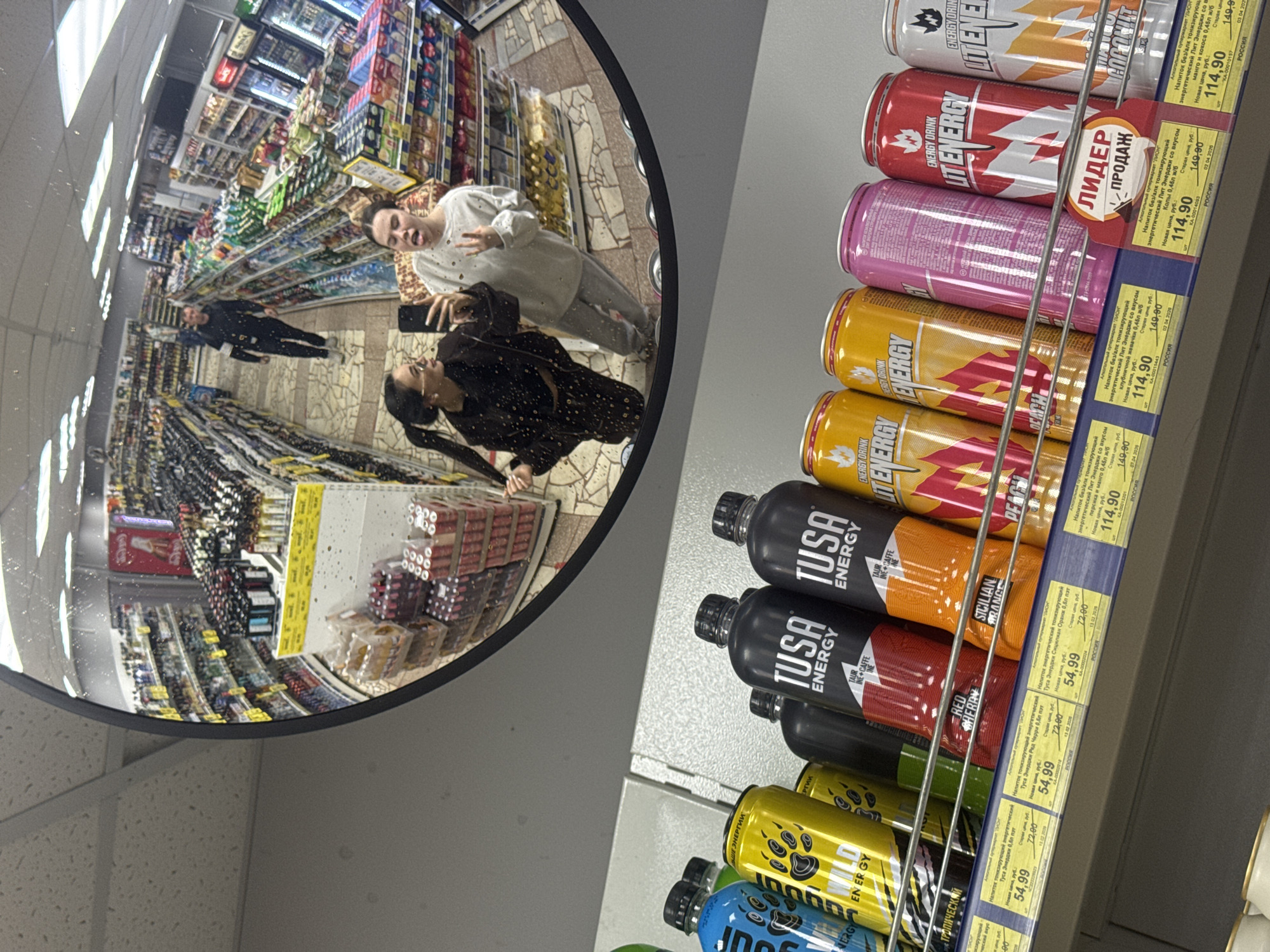The image shows a shelf of energy drinks in a store, as reflected in a convex mirror. The drinks include brands like "Lit Energy" and "TUSA Energy" with various flavors, each with price tags in Russian rubles. The reflection in the mirror reveals a supermarket aisle with shoppers. Two people, one holding a phone, are reflected in the mirror, taking a picture of themselves with the shelf. The image suggests the location is a supermarket, likely in Chaikovsky, Russia, based on the currency and text on the price tags.