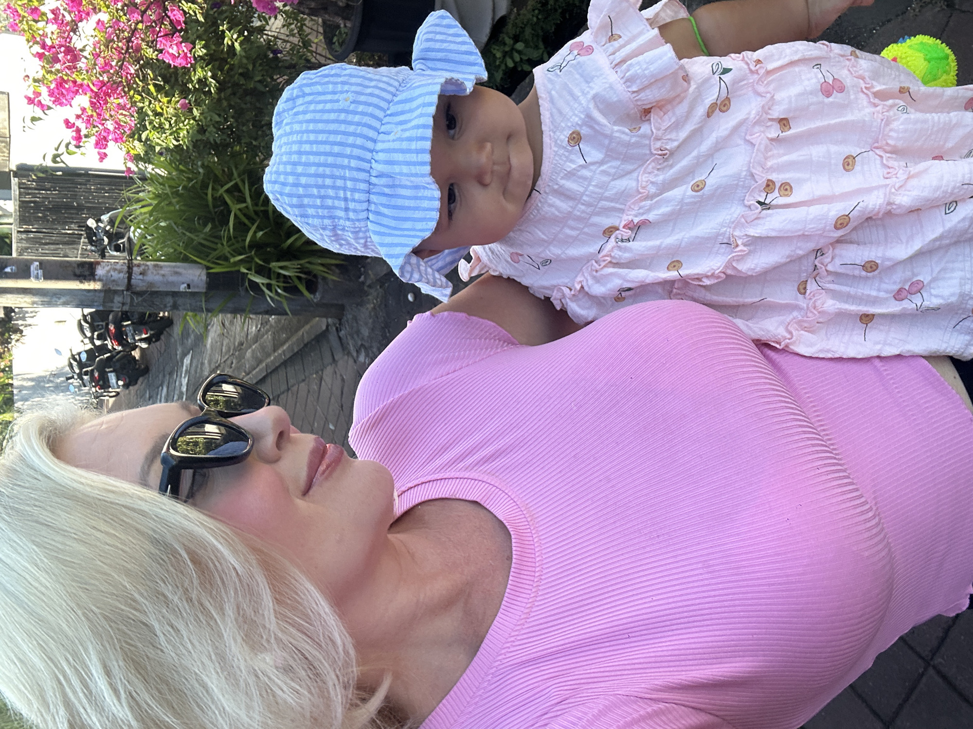 A woman with platinum blonde hair and black sunglasses is holding a baby, presumably her child, in North Kuta, Indonesia. The woman is wearing a pink, ribbed top. The baby is wearing a white and pink dress with a cherry pattern and a light blue and white striped sunhat. In the background, there are potted flowers, a utility pole, and motorcycles parked along the street. The scene is brightly lit, suggesting it is daytime.