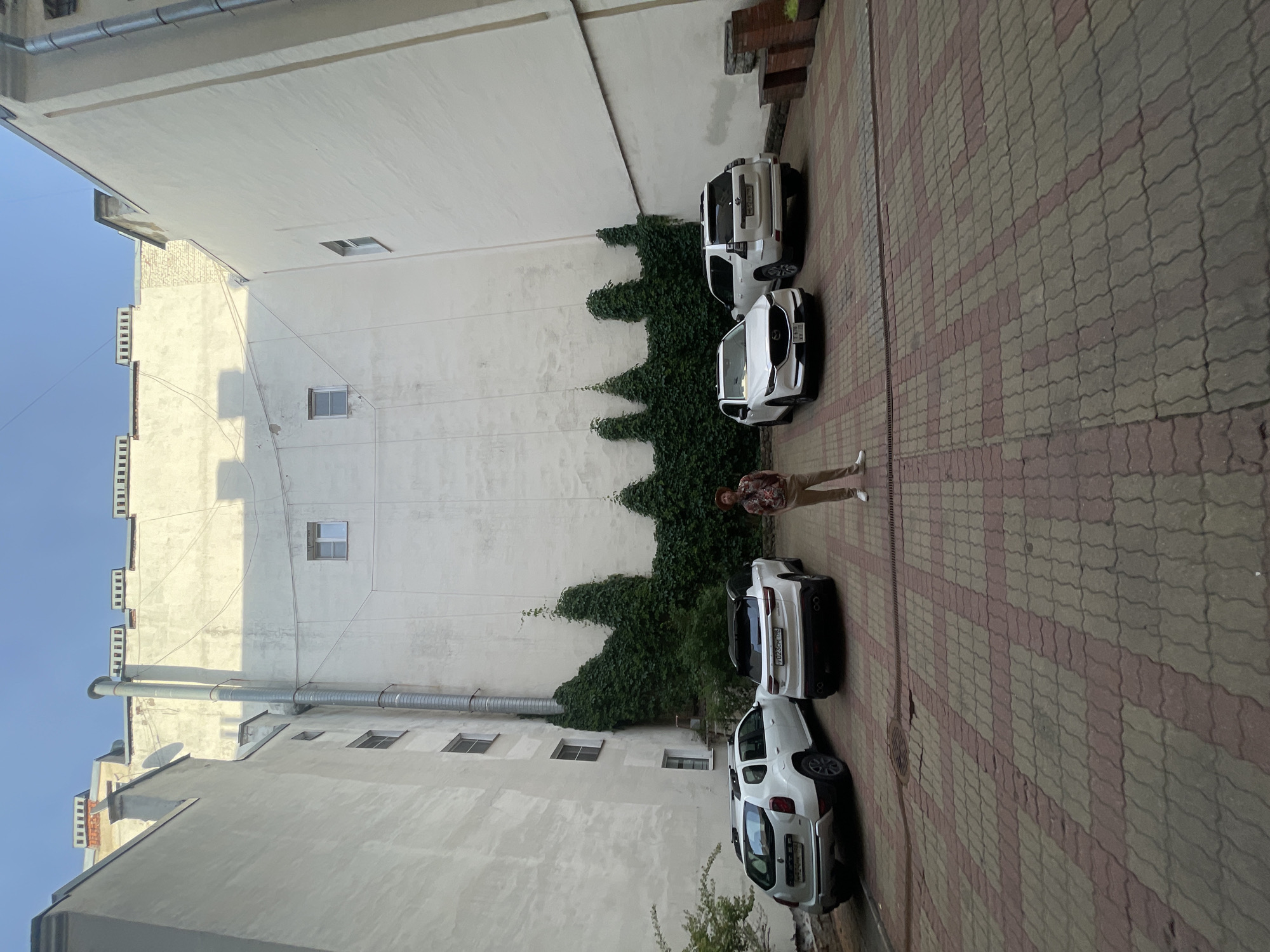 The image depicts a courtyard in Saint Petersburg, Russia, with tall buildings on three sides. The courtyard is paved with red and grey interlocking bricks. In the foreground, a man is standing to the right of the frame. He is wearing a patterned shirt, light brown pants, and a straw hat. There are four cars parked in the courtyard. From left to right, they are a white Dacia Duster, a white crossover SUV, a white Mazda CX-5, and a white Toyota Land Cruiser Prado. Lush green ivy grows up the side of the buildings, creating decorative patterns. The sky is a clear blue with a slight haze. The overall scene suggests an urban environment, possibly residential or commercial, with a focus on parking and architecture.