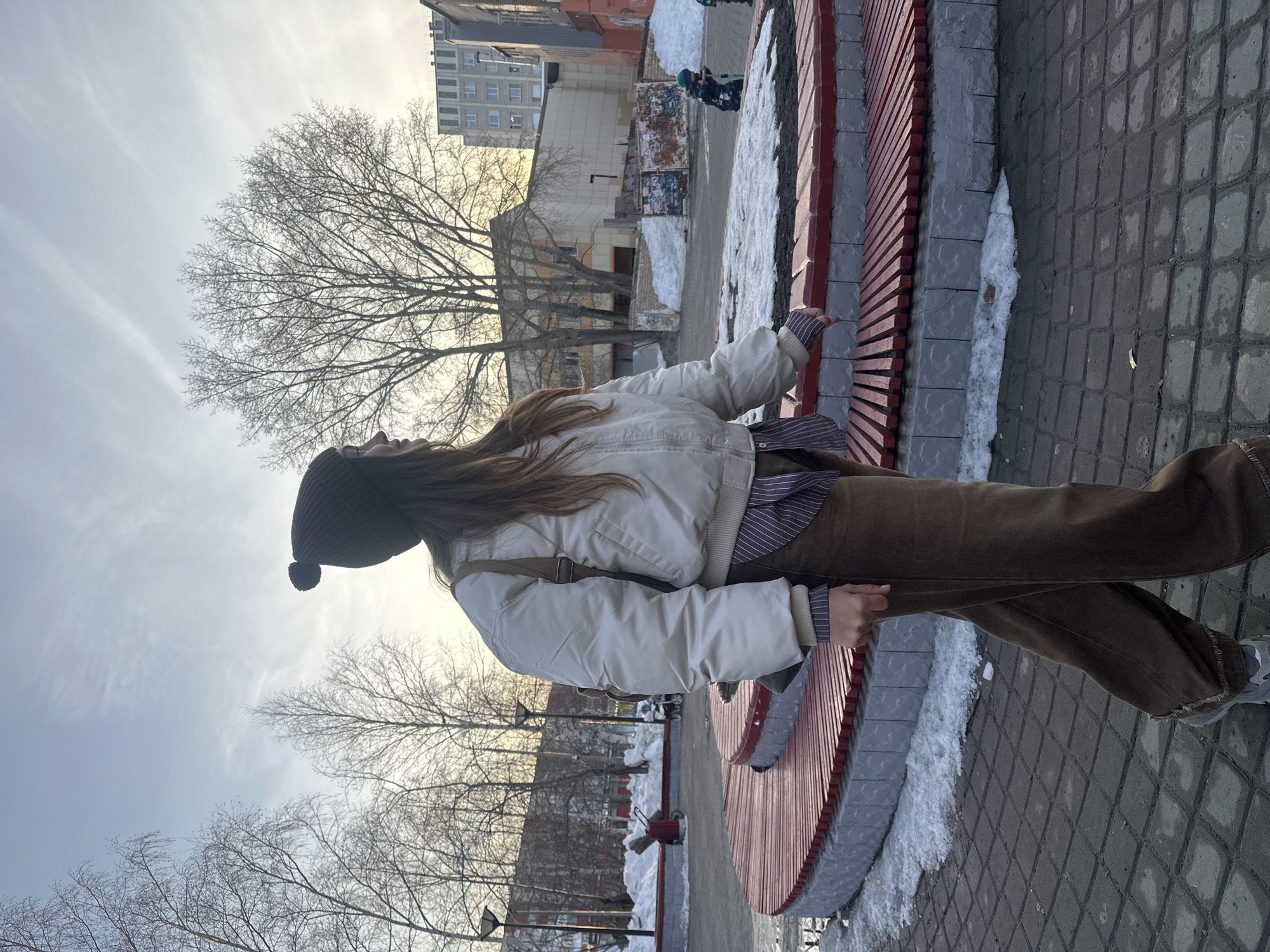A woman stands on a brick-paved area next to a circular red and gray bench. She wears a dark beanie, a white jacket over a striped shirt, and brown pants. The woman looks upwards. Behind her are buildings, trees, and patches of snow. Two children are visible in the background. The location is likely Surgut, Russia.