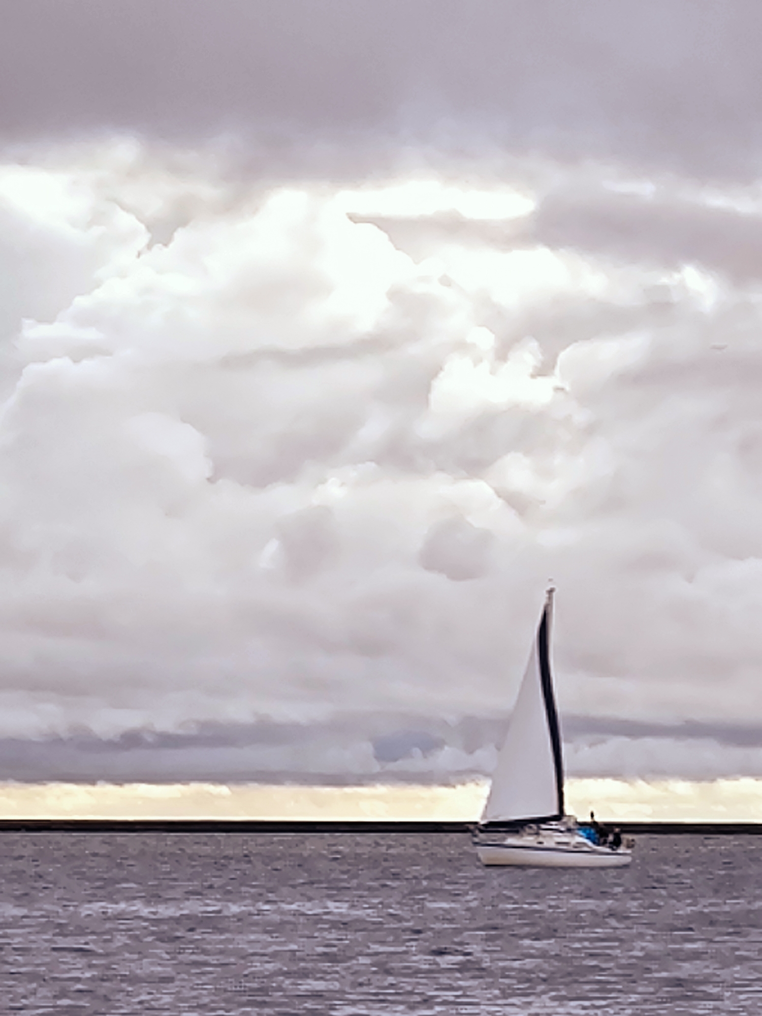 An image captures a white sailboat with black trim sailing on a dark, rippling body of water under a dramatic sky filled with light and dark grey clouds. A thin, bright horizontal line of light is visible on the horizon between the water and the clouds. The sailboat, positioned in the right half of the frame, has its main sail up, catching the light. Figures are visible on the deck of the boat, but their details are indistinct. The overall atmosphere of the image is somber and serene, with the interplay of light and shadow on the clouds creating a sense of depth and mood. The scene is characteristic of coastal areas, potentially along the Daugava River in Riga, Latvia, as it meets the Baltic Sea.