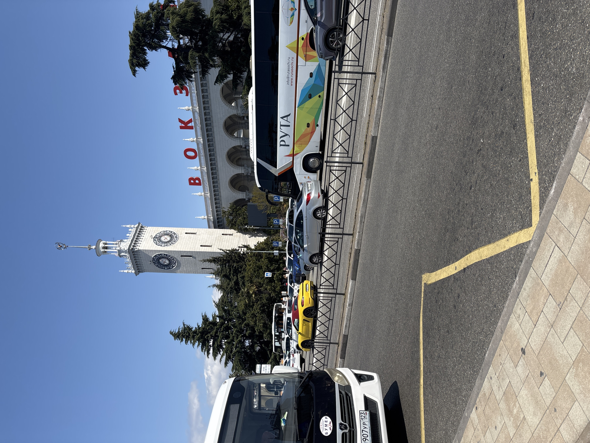 The image shows a view of Sochi, Russia, featuring the Sochi Sea Port building prominently. The building is white with arched windows and a clock tower. The word "ВОКЗАЛ" is displayed in red on the building's facade. A line of vehicles, including buses and cars, is parked along the street next to a metal railing. The street is marked with a yellow line along the curb. Trees are visible, framing the building in the background. The sky is clear and blue. The scene appears to be a busy area with transportation services.