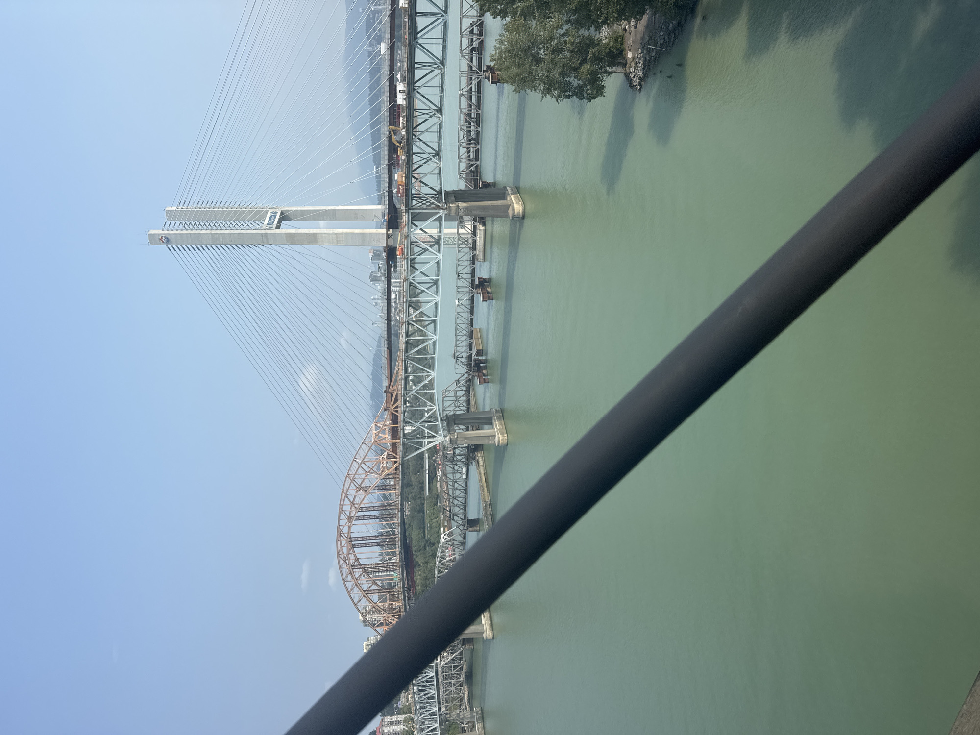 The image depicts a wide shot of a body of water, likely a river, with several bridges spanning across it. In the foreground, a dark, thick railing or bar is diagonally positioned, partially obscuring the view.
The most prominent structure is a large, modern cable-stayed bridge with two tall, white concrete towers. Numerous cables extend from the towers to the bridge deck. To the left of this bridge, an older, orange-colored truss bridge with arched supports is visible. Further to the left, another gray truss bridge with visible structural elements can be seen. In the background, more bridges and a city skyline with buildings are apparent, along with a distant mountainous landscape under a clear blue sky with a few wispy clouds.
On the far right side of the image, a section of the riverbank is visible with trees and rocks. The water itself is a greenish hue, reflecting the sunlight. The overall scene suggests an urban environment with significant infrastructure, likely in a location with a significant river.
Based on the presence of multiple bridges, including the Alex Fraser Bridge and the Pattullo Bridge, and the general topography, this scene is likely located in Surrey, Canada, specifically in the vicinity of the Fraser River. The bridges are iconic landmarks of the region, facilitating transportation across the water. The image captures a sunny day with good visibility. There are no people or animals clearly visible in the scene.
