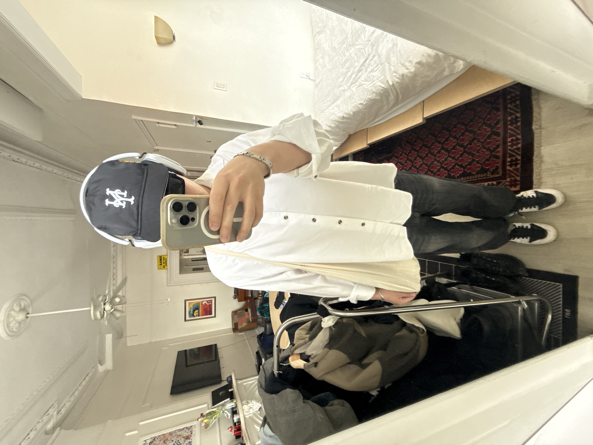 A person is taking a selfie in a mirror. The person is wearing a white button-down shirt, dark jeans, and black and white sneakers. They are also wearing a New York Mets baseball cap. The background of the photo includes a bed, a rug, a clothing rack, and other items. The location appears to be an apartment or residential setting in New York City, United States.