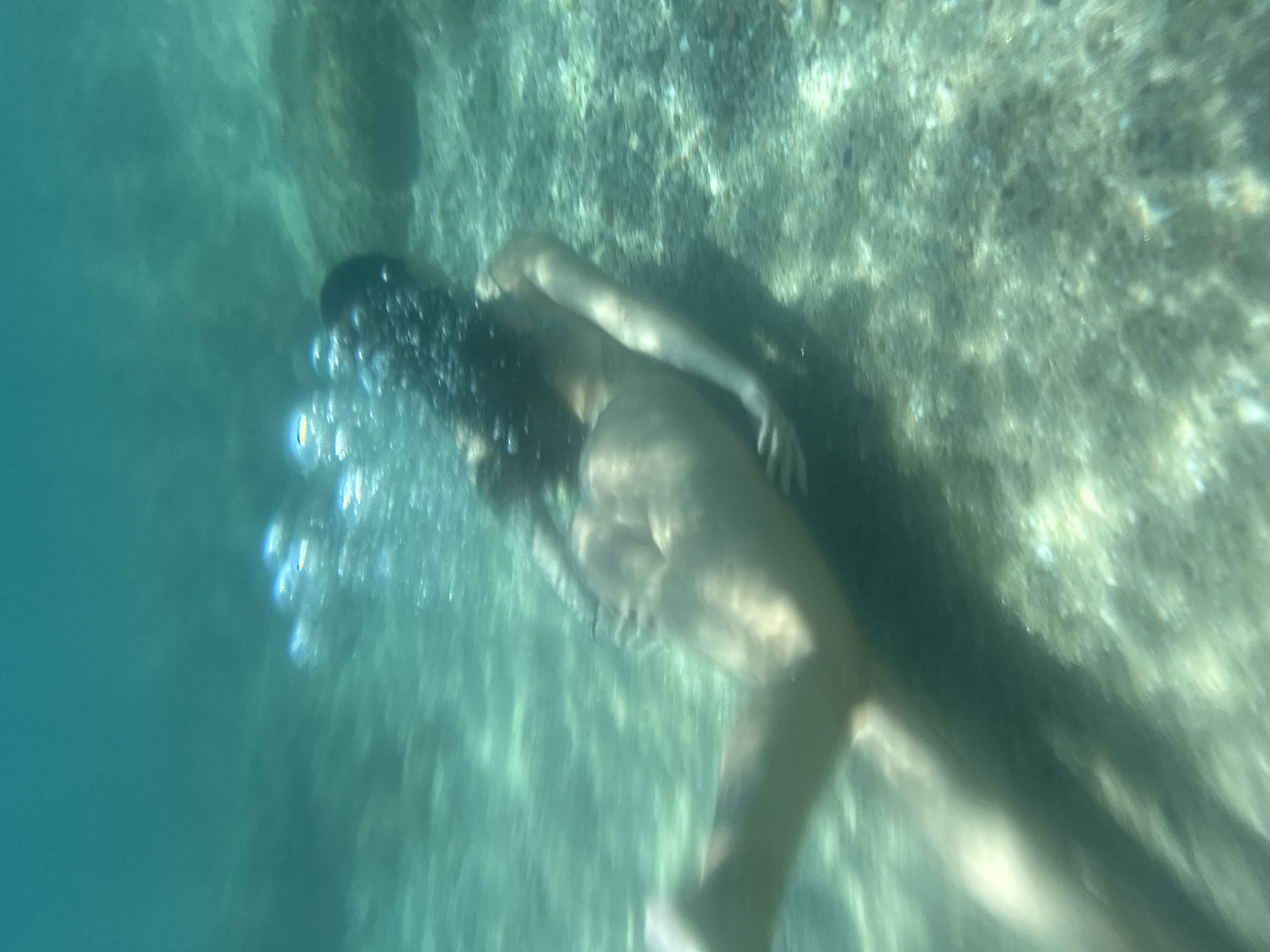 This is an underwater photograph taken in the clear, turquoise waters off the coast of Arenys de Mar, Spain. The scene depicts a person swimming. The individual has dark hair and appears to be moving away from the camera, with their head and upper body visible. Bubbles are rising around them, indicating they are either exhaling or have just surfaced from a dive. The lighting suggests it's daytime, with sunlight filtering through the water. The background is a mix of natural underwater elements, including what appears to be a rocky seabed or reef with some vegetation. The overall impression is one of serene, natural movement within an aquatic environment. There is no visible text in the image.