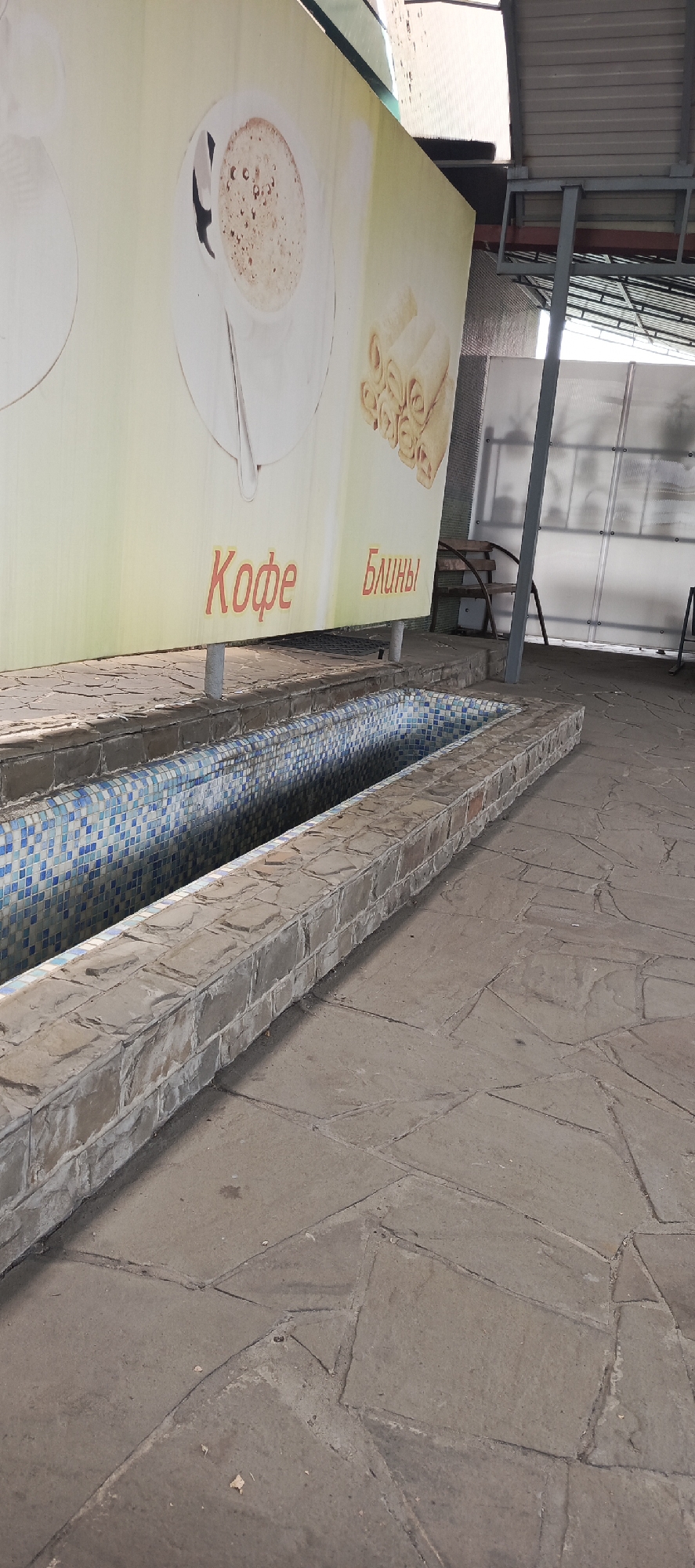 The image shows an outdoor area with a tiled trough or water feature in the foreground. The trough is rectangular and filled with blue and white tiles. The surrounding area is paved with large, irregular stone slabs. Behind the trough, a large banner displays images of coffee and rolled pancakes, with Russian text that translates to "Coffee" and "Pancakes". In the background, there is a covered seating area with a metal structure and translucent panels, and a wooden bench. The scene appears to be located in Kamenolomni, Russia, possibly an outdoor cafe or rest area. There are no people visible in the image.