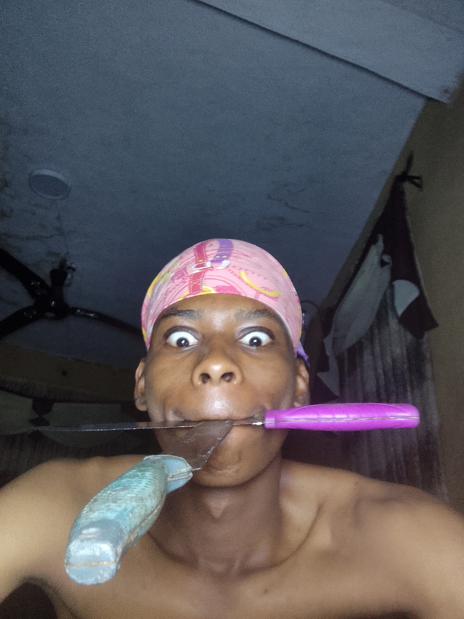 The image features a person with dark skin and large, wide eyes, wearing a pink headscarf with various cartoon images. They are holding two knives in their mouth, one with a purple handle and the other with a blue handle. The person appears shirtless. Behind them, the background consists of a ceiling with a light fixture and a ceiling fan on the left. There's a draped garment hanging on the right. The location is in a room in the city of Unknown, Unknown.