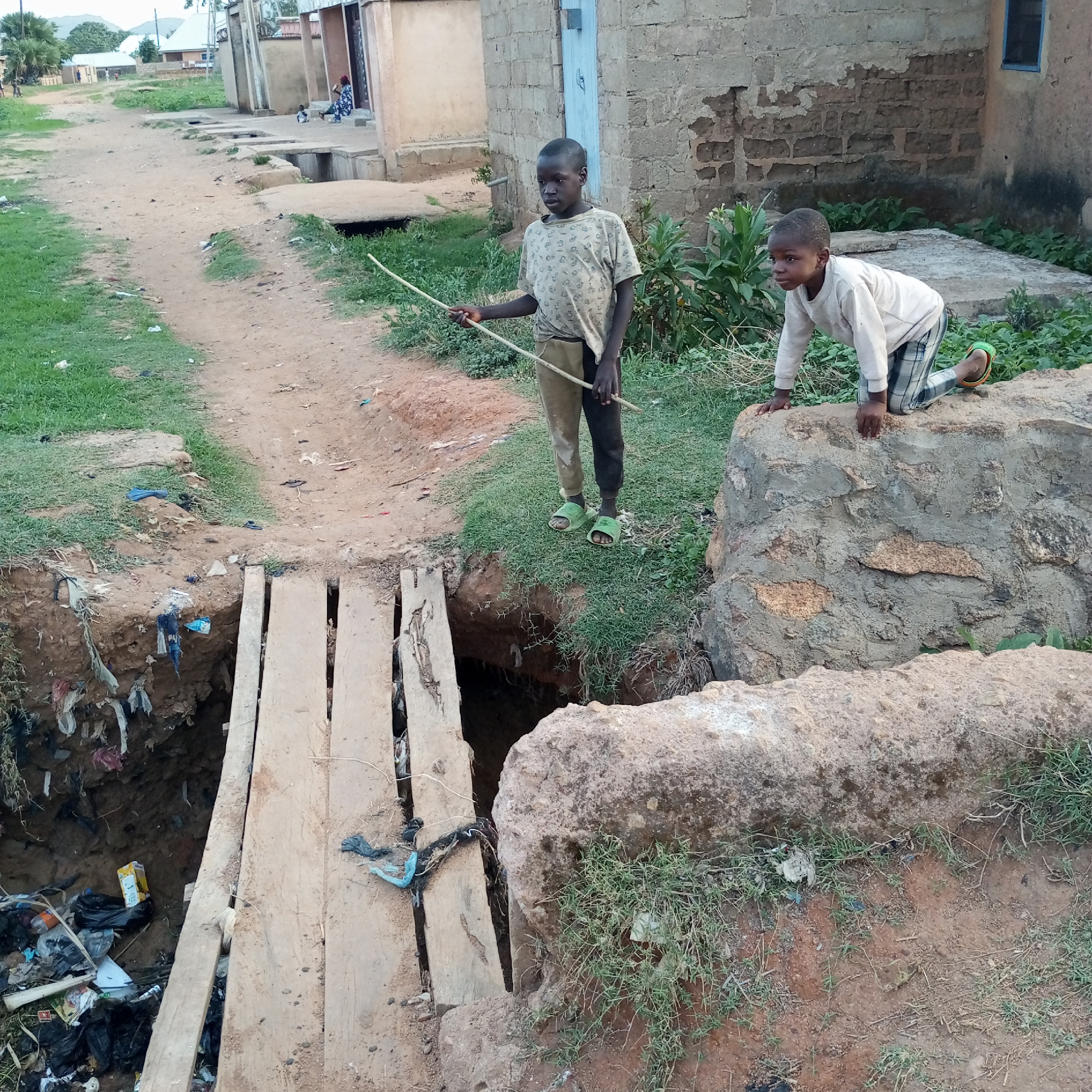 Two young boys are in Bukuru, Nigeria, near a large trench that spans a dirt road. One boy stands at the edge of the trench with a stick in his hand, while the other is climbing on a large stone. A makeshift bridge constructed of wooden planks spans the trench. The trench is filled with garbage. Buildings are visible in the background.