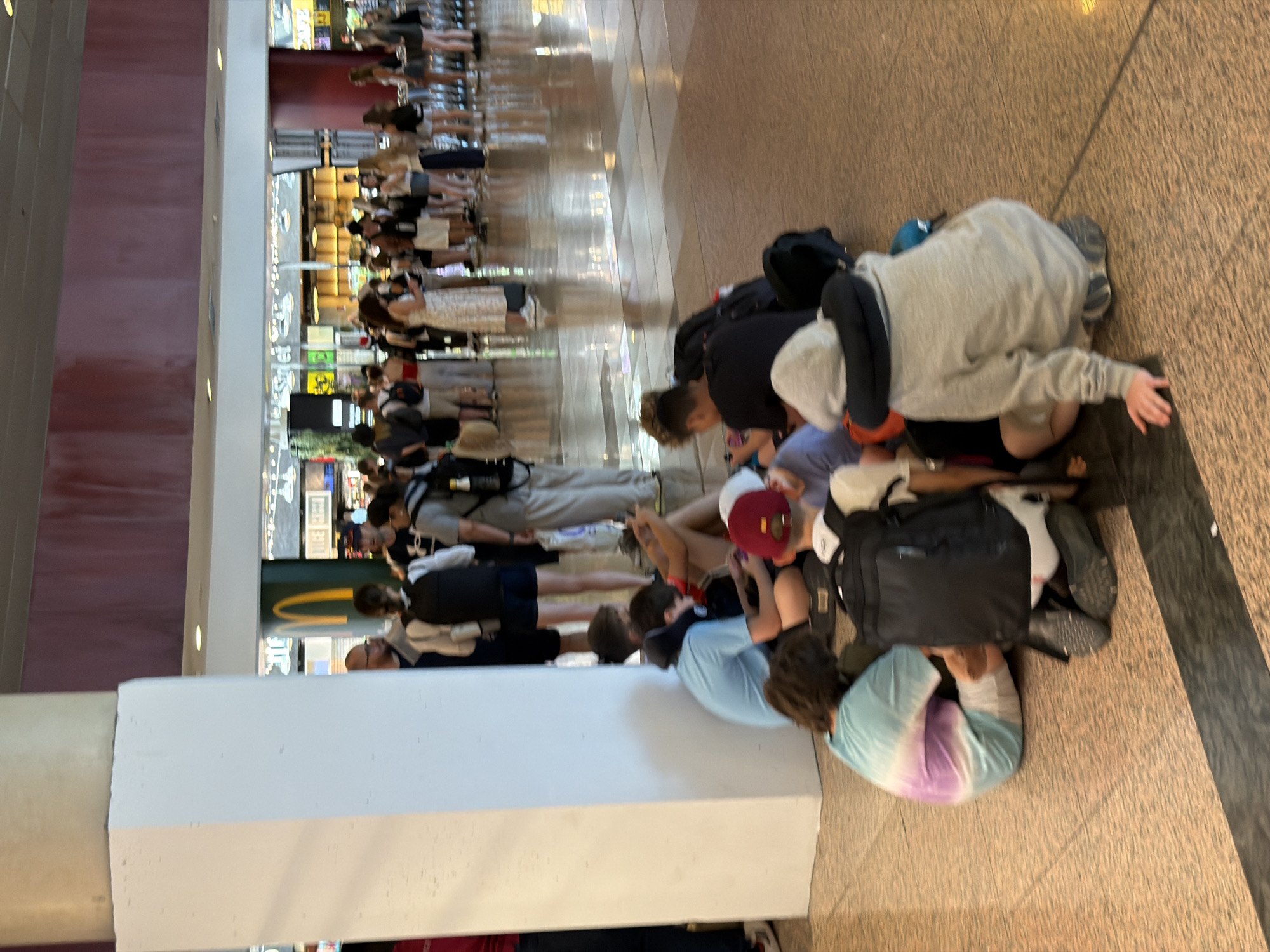 The image depicts a group of people, mostly young individuals, sitting on the floor in what appears to be a public transit hub or waiting area. In the foreground, several individuals are seated on the ground, some with backpacks. One person is wearing a tie-dye t-shirt, another a grey hoodie with a neck pillow. In the background, more people are standing, some in a queue. The environment features polished floors, a white pillar, and the visible signage of a McDonald's restaurant, suggesting a commercial area within an airport or a large shopping center. The overall scene conveys a sense of waiting or resting in a busy public space. Given the presence of McDonald's and the general ambiance, this location is likely within the Barcelona-El Prat Airport (BCN) in El Prat de Llobregat, Spain, a common area for travelers.