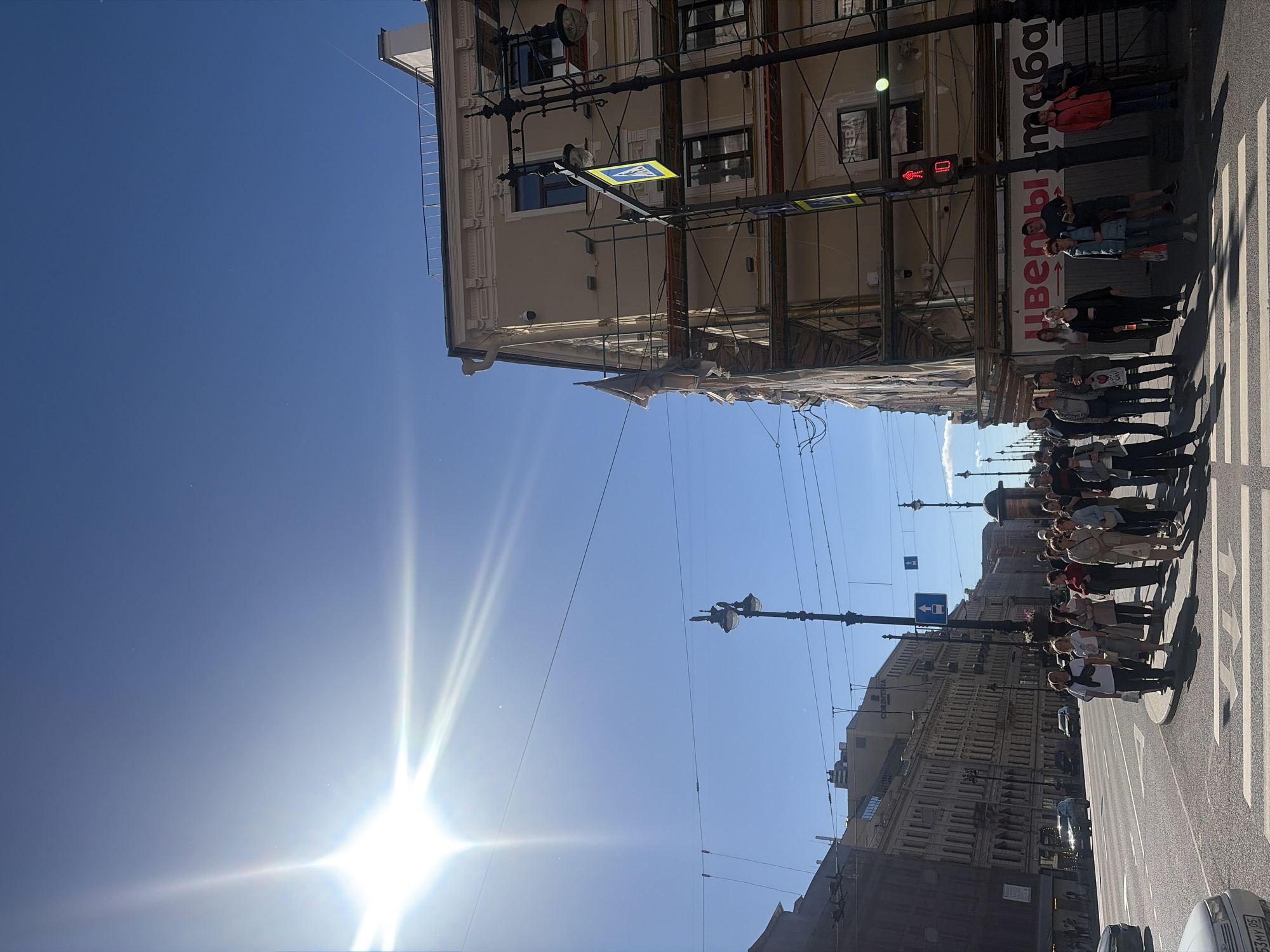 The image depicts a sunny day scene on a street in Saint Petersburg, Russia. The bright sun is visible in the upper left corner, creating lens flare. The street is wide, with multiple lanes of traffic and a pedestrian crossing. Buildings line the street, some appearing to be undergoing renovation with scaffolding. Several people are standing on the sidewalk or at the pedestrian crossing, waiting to cross the street. There are streetlights, traffic lights, and traffic signs visible. The architecture suggests a historic European city. The overall impression is a bustling urban environment under a clear blue sky.
