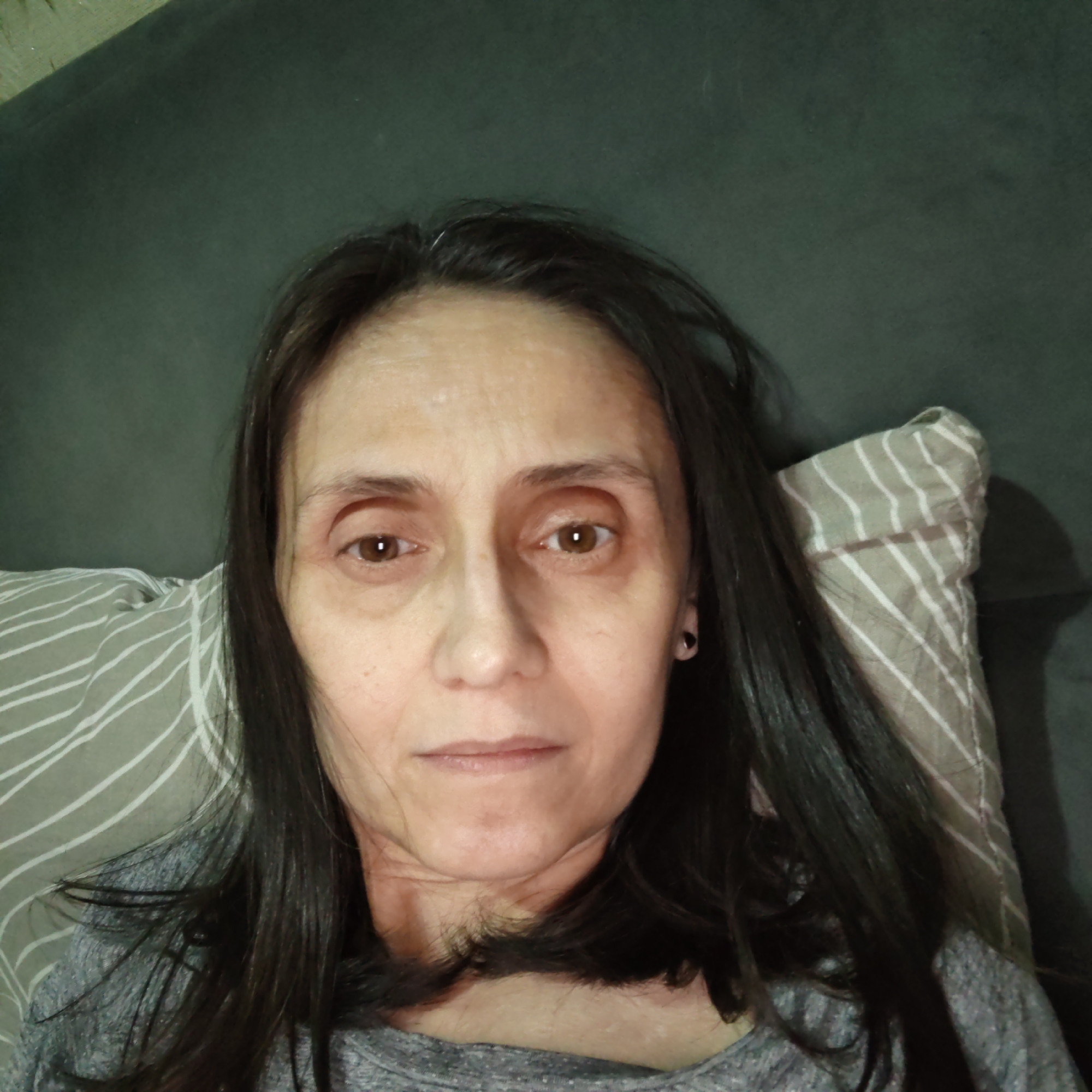 A medium shot shows a woman lying down, looking directly at the camera. She has long, dark hair and is wearing a gray shirt. The background is a dark green couch with a pillow that has a striped pattern. The lighting is dim, creating shadows on her face. There is a pillow behind her head, partially visible on the right side of the frame. The woman has a black stud earring in her visible ear.