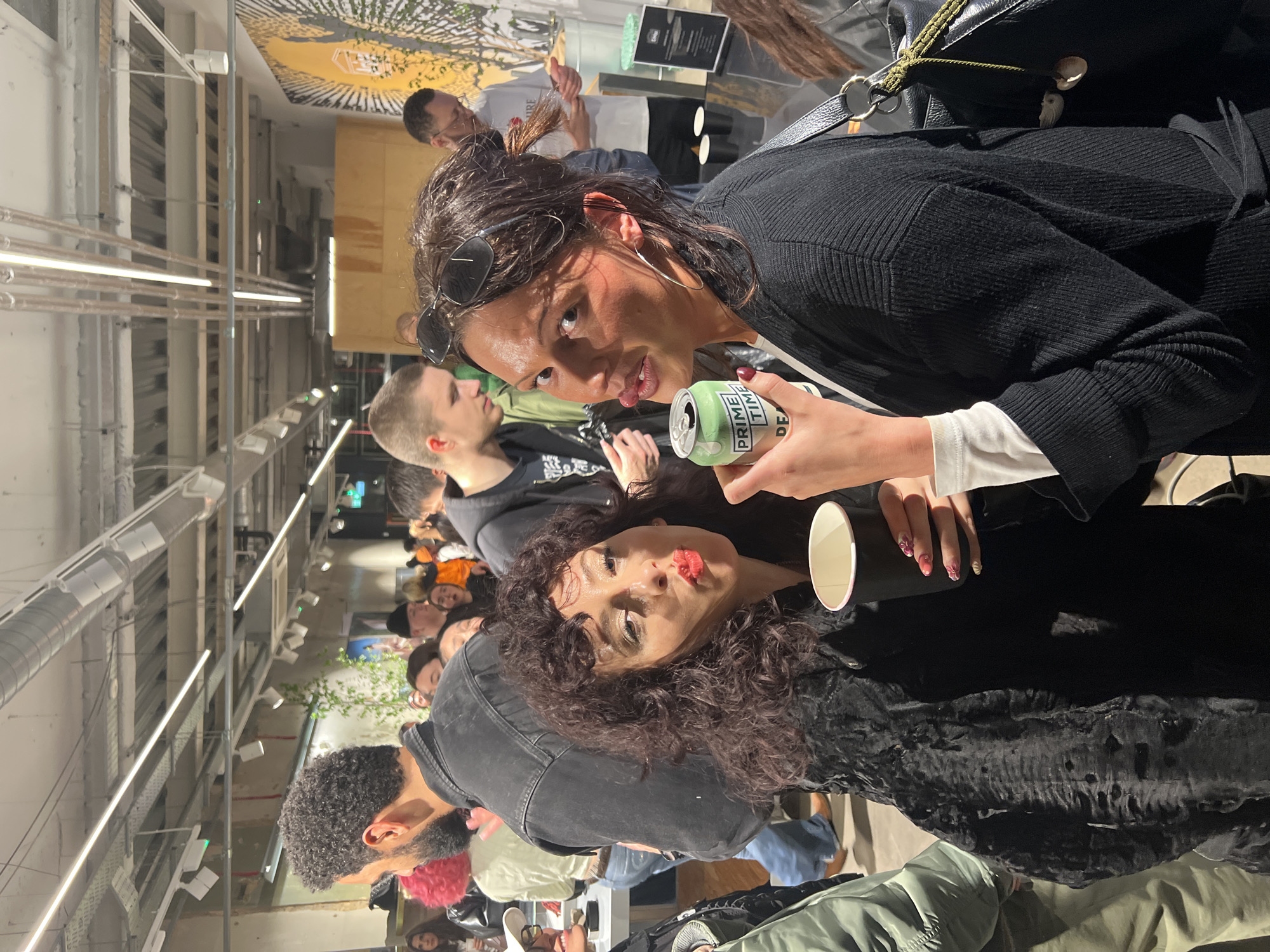 The image depicts an indoor social gathering or event, bustling with numerous young adults. Two women are prominent in the foreground, posing directly for the camera. The woman on the right has dark hair pulled back with sunglasses on her head, wearing a black cardigan over a white top. She holds a green can, clearly labeled "PRIME TIME" with "DE" also visible, and looks at the camera with a playful expression. A black handbag with a green strap is slung over her shoulder.
To her left, another woman with dark, curly hair and bright red lipstick looks at the camera with pursed lips. She holds a black paper cup and a small, round mirror. Both women appear engaged and lively.
The background is filled with other attendees, many blurred or partially obscured, suggesting a crowded environment. Notable details among them include a man with curly hair and a beard, and another individual with vibrant pink/red hair. The setting features an industrial aesthetic with an exposed ceiling revealing pipes, conduits, and numerous long, linear fluorescent light fixtures. A large, colorful mural or graphic, possibly depicting a tree, adorns a back wall. A black display stand with illegible text is also visible further in the background. The scene suggests a casual, perhaps evening, social event.