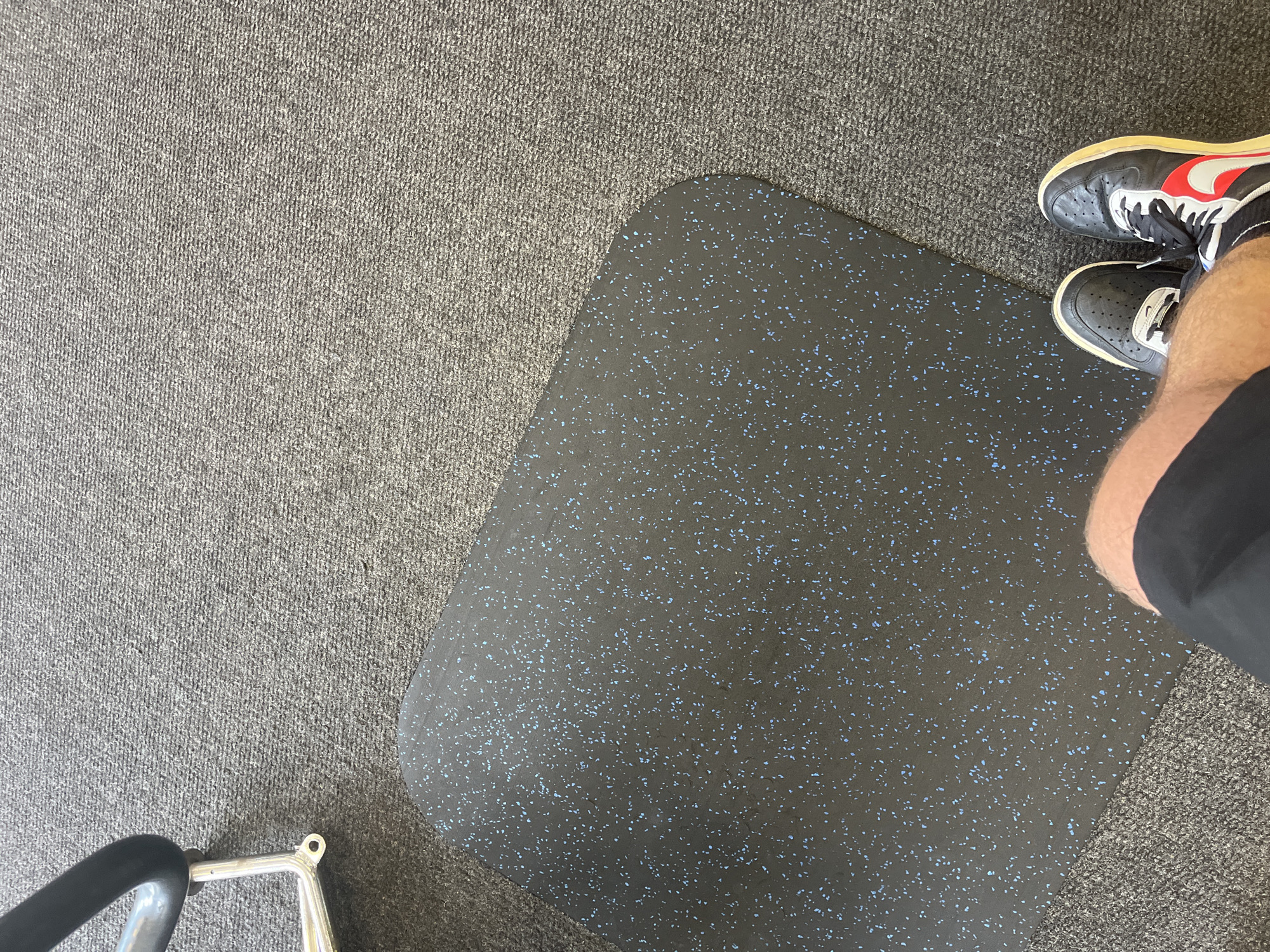 An overhead shot captures a person's lower legs and feet within an indoor setting, likely a gym or workout area, in Blagoevgrad, Bulgaria. The person is wearing black shorts and black sneakers with white soles and red and white accents, possibly Nike, with one shoe clearly showing a red swoosh.
The floor is divided between two main surfaces: a light gray, textured carpet with a short pile covering the left and upper parts of the frame, and a dark gray rubber mat with numerous small, light blue flecks occupying the central and right areas. The rubber mat features rounded corners and a smooth surface.
Part of a metallic piece of exercise equipment, including a silver frame and a black padded handle, is visible in the bottom left corner of the image. The lighting suggests an interior space. The person appears to be standing still, and no specific activity is taking place. There is no discernible text visible on any objects or surfaces.