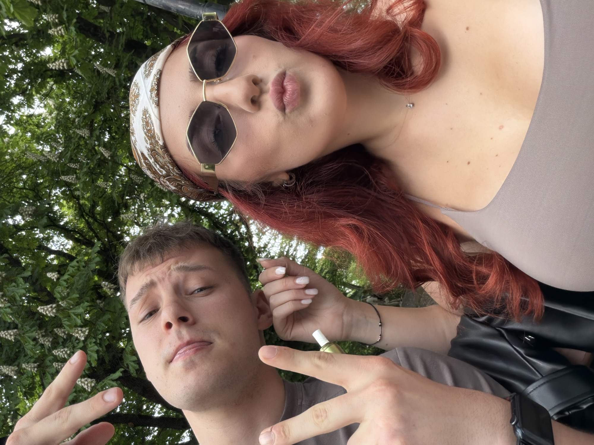 Two people pose in a selfie. The individual in the foreground is a young man, with short, light-brown hair. He is making a peace sign with his fingers. Behind him is a woman with reddish-auburn hair, sunglasses, and a headscarf. She is wearing a fitted top and a necklace. She is pursing her lips. The background includes foliage and white blossoms, suggesting an outdoor location. The scene is likely in Sofia, Bulgaria.