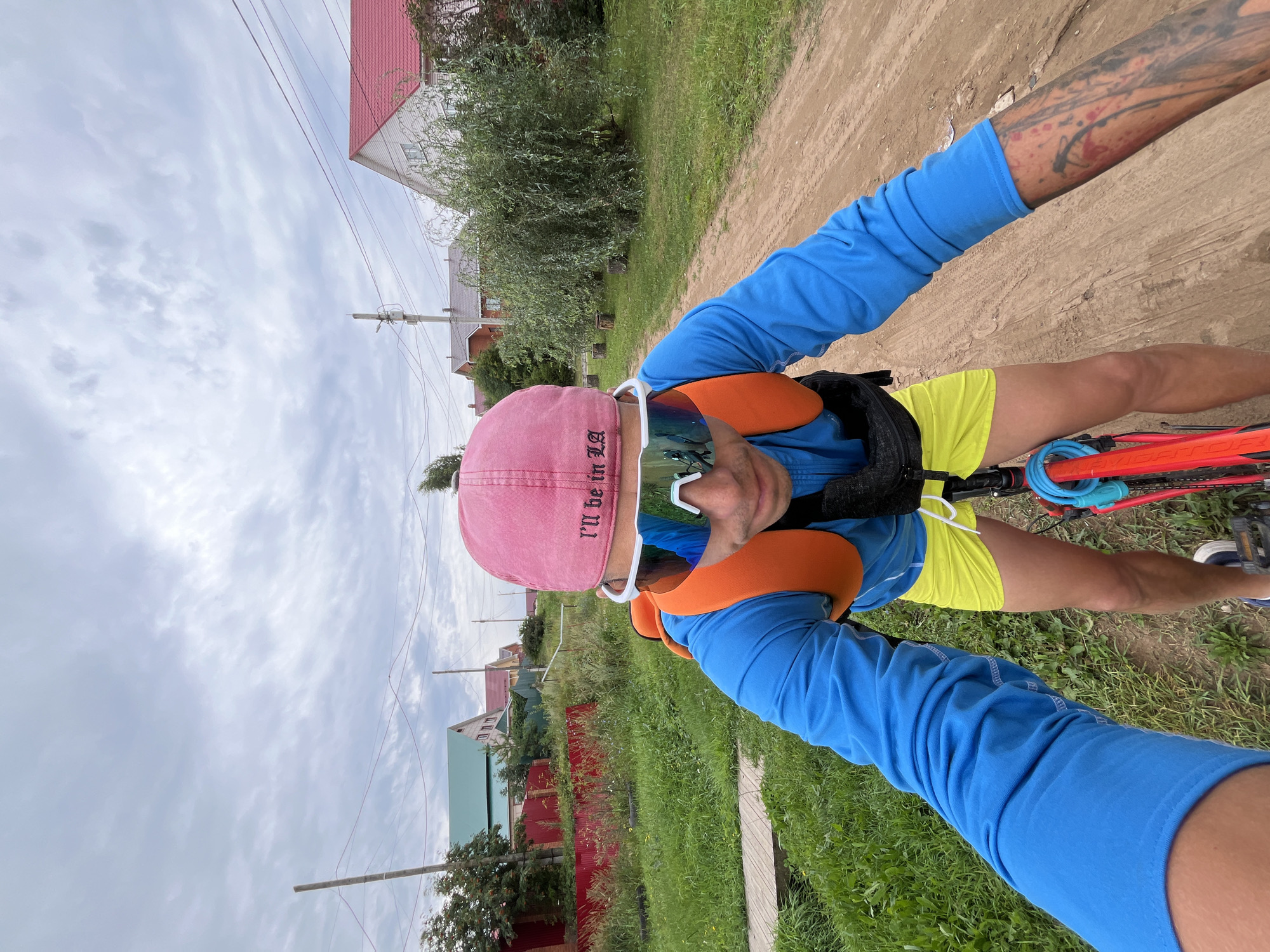 A person is riding a bicycle on a dirt road in a rural area. The person is wearing a blue long-sleeved shirt, yellow shorts, a pink baseball cap, and white cycling glasses. The person has an orange backpack. There are houses with red roofs in the background, along with trees and grass. The sky is cloudy. The image was taken in the city of Chaikovsky, Russia.