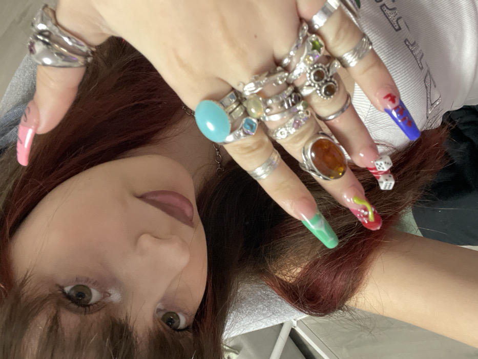 A woman with brown hair gazes upwards, showcasing a multitude of rings on her fingers. The rings range in style, from chunky silver bands to those adorned with colorful gemstones like turquoise and amber. Her nails are long and painted in a variety of vibrant colors and designs, including dice. She wears pink lipstick and appears to be indoors.