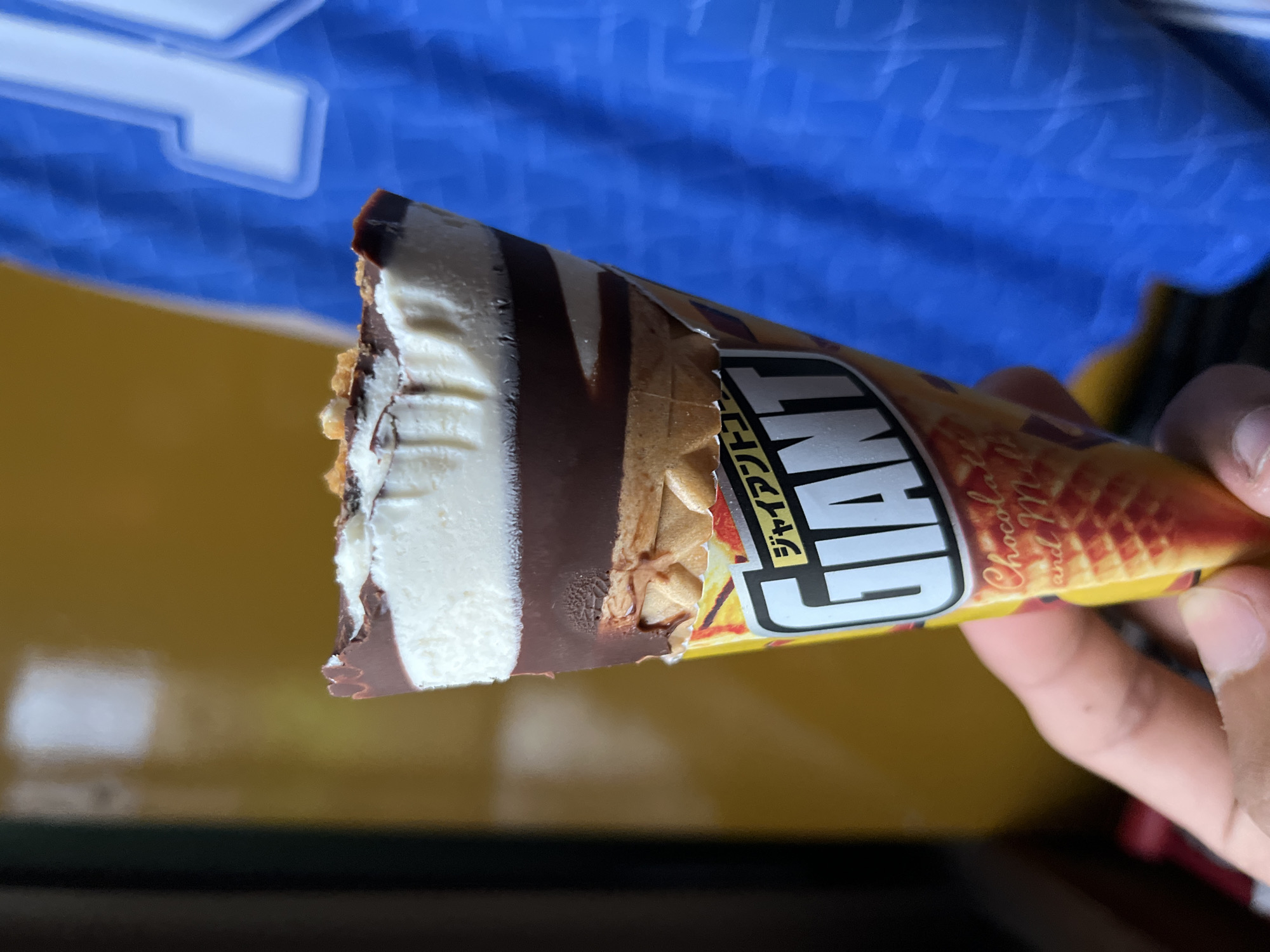 An ice cream cone, possibly a Giant brand, is held by a person's hand in front of a yellow and blue background. The ice cream has a white top layer, a dark brown chocolate coating, and a tan waffle cone. Text on the cone's wrapper appears to be in Japanese and English, with "GIANT" prominently displayed. The background suggests an indoor setting, possibly a home or shop, with a yellow wall on the left and a blurred blue fabric, perhaps clothing or a banner, on the right. The image appears to be taken in Sayama, Japan.