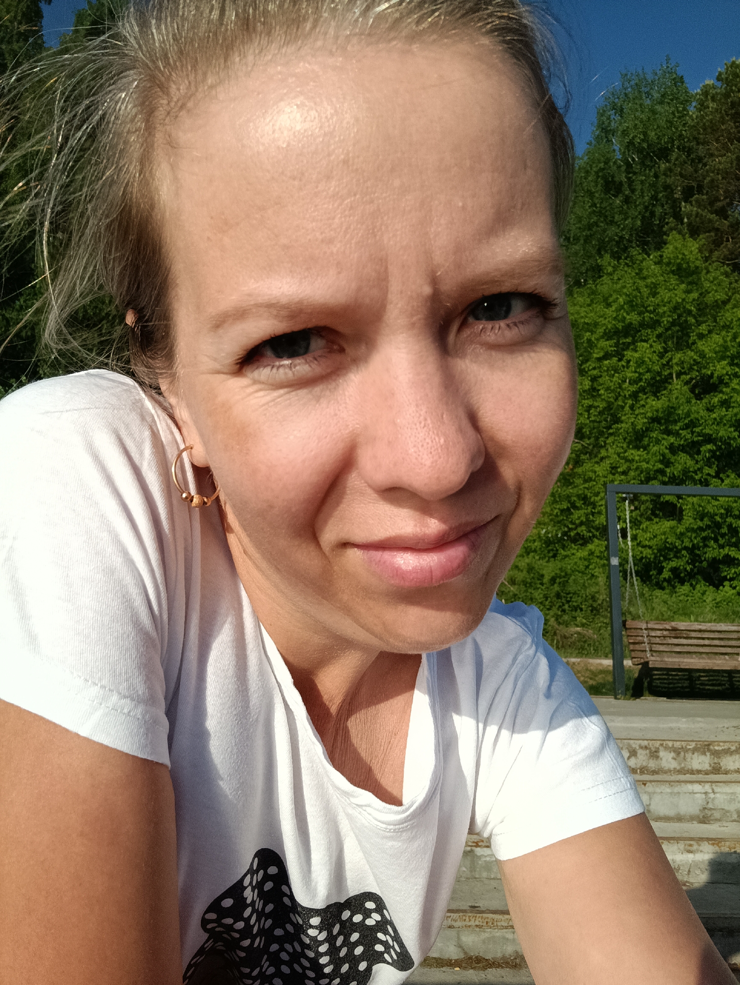 A close-up selfie shows a fair-skinned woman with blonde hair pulled back, wearing a white t-shirt with a black and white pattern near the bottom. She has light eyes and a slight smirk. In her left ear is a gold hoop earring. The background includes green trees, a patch of blue sky, and a wooden park bench swing. The photo was taken outdoors on a sunny day. The exact location of the park in the city of Chaikovsky, Russia is not discernible from the image.