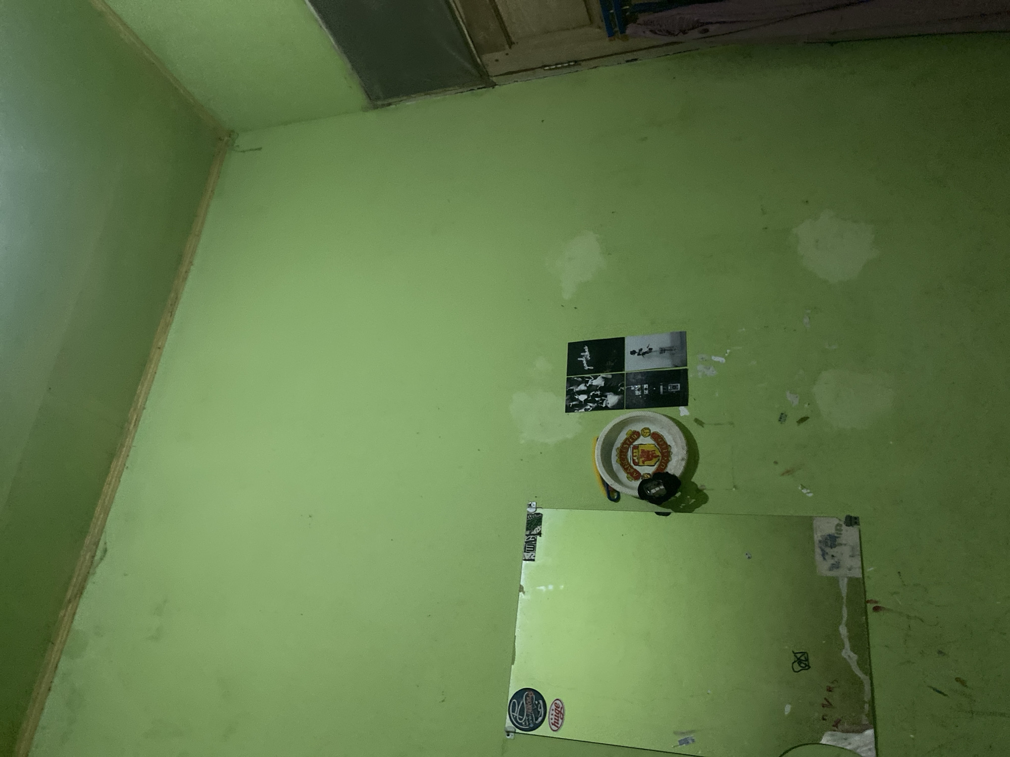 The image depicts an interior corner of a room with light green walls and ceiling. The lighting is dim and appears to be artificial, casting shadows. On the left side of the image, a portion of a large, light green rectangular object, possibly a whiteboard or a mirror, is visible. It is adorned with several stickers, including one oval-shaped sticker with the word "huge" at the bottom and a circular sticker above it featuring a blue logo with white text. To the right of this object, there is a small, circular dish with the Manchester United crest, a football club logo, displayed on it. A black object, possibly a flashlight or a remote, is attached to the dish. Further to the right, on the wall, are several small, black and white posters arranged in a grid-like fashion. One poster appears to be a collage of people in a performance, while another shows a person standing. The walls show signs of wear and tear, with several irregular white patches and some dark marks. To the far right, a portion of a wooden door frame and an open doorway revealing a dark interior with some fabric hanging can be seen. The overall scene suggests a personal space, possibly a bedroom or a study, in a dwelling in Ta', Indonesia.