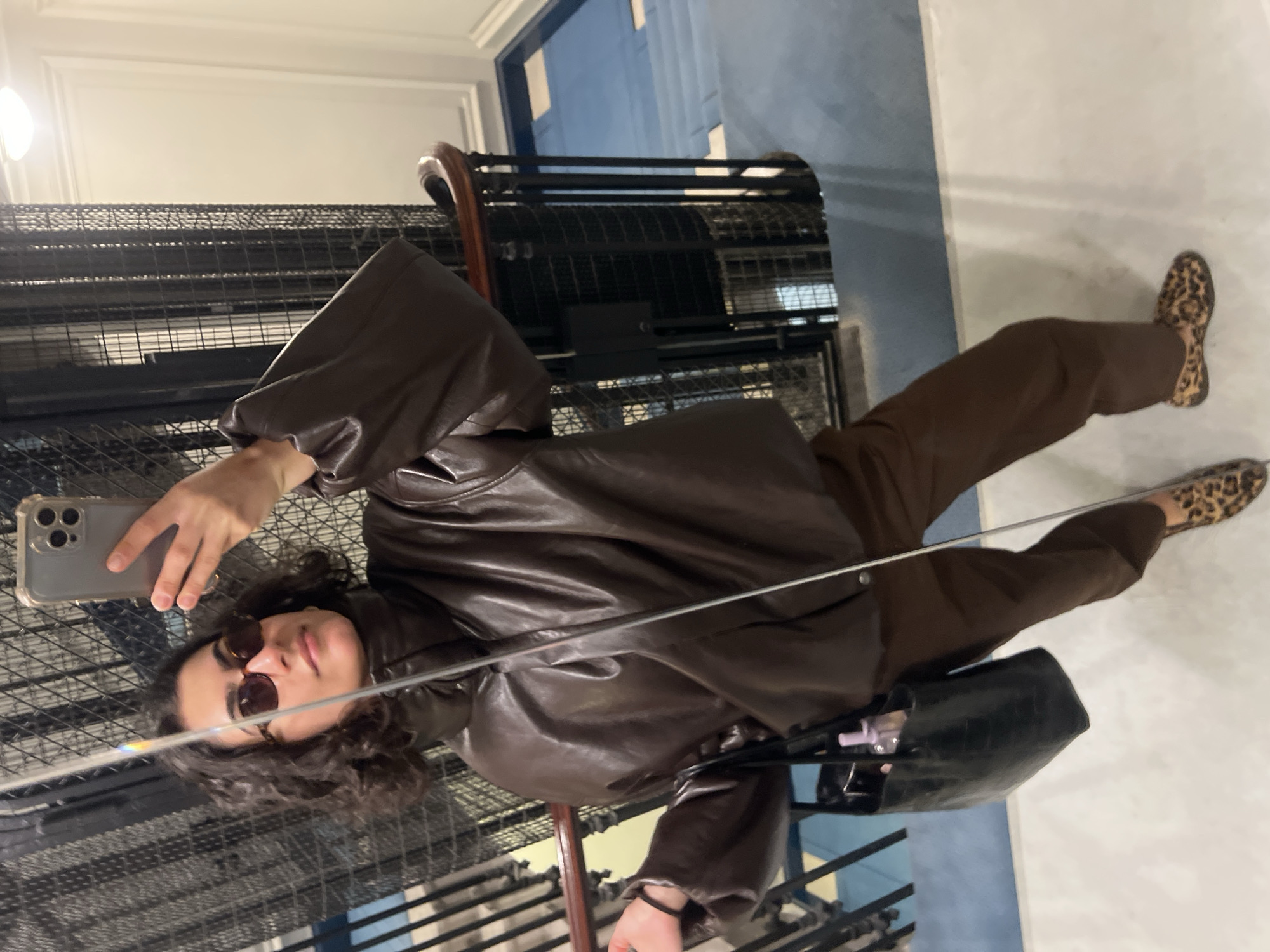 A person is reflected in a mirror located on a stairway with black metal railings. The person is holding a smartphone in their right hand, appearing to take a selfie. They are wearing sunglasses, a brown leather jacket, and brown trousers. On their feet are animal print loafers. A black handbag hangs off the person's left shoulder. The stairs are located indoors with blue and white walls. The location of the stairway is in Paris, France.