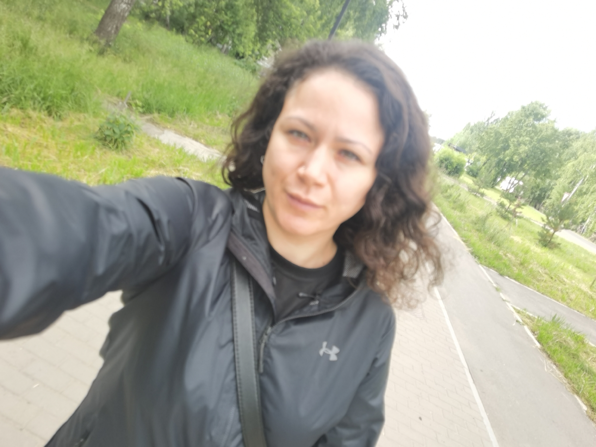 A woman with curly brown hair is taking a selfie outdoors. She is wearing a black hooded jacket with a zipper and an Under Armour logo on the chest, and a black t-shirt underneath. She has a dark gray crossbody bag slung over her left shoulder. The background shows a paved walkway next to a grassy area with trees. The scene appears to be a park or a residential area. The lighting is bright and overcast. The location is Chaikovsky, Russia.