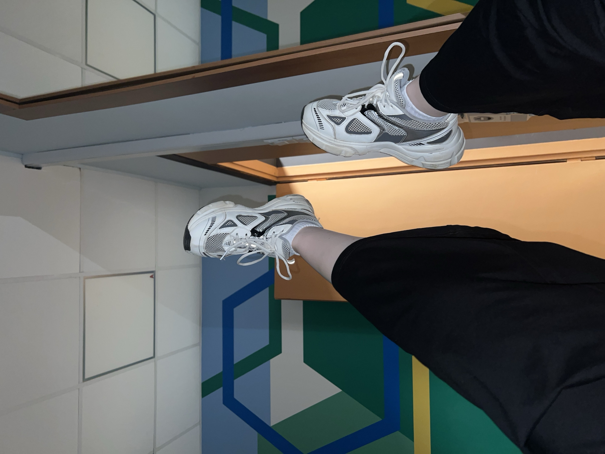The image shows a pair of legs wearing black pants and white sneakers against a geometrically patterned wall. The legs are positioned upside down, suggesting the perspective is from a mirror or an inverted camera angle. The background features a pattern of interconnected polygons in shades of blue, green, and white. The location in Khimki, Russia, is not identifiable based solely on the visual elements present in the image.