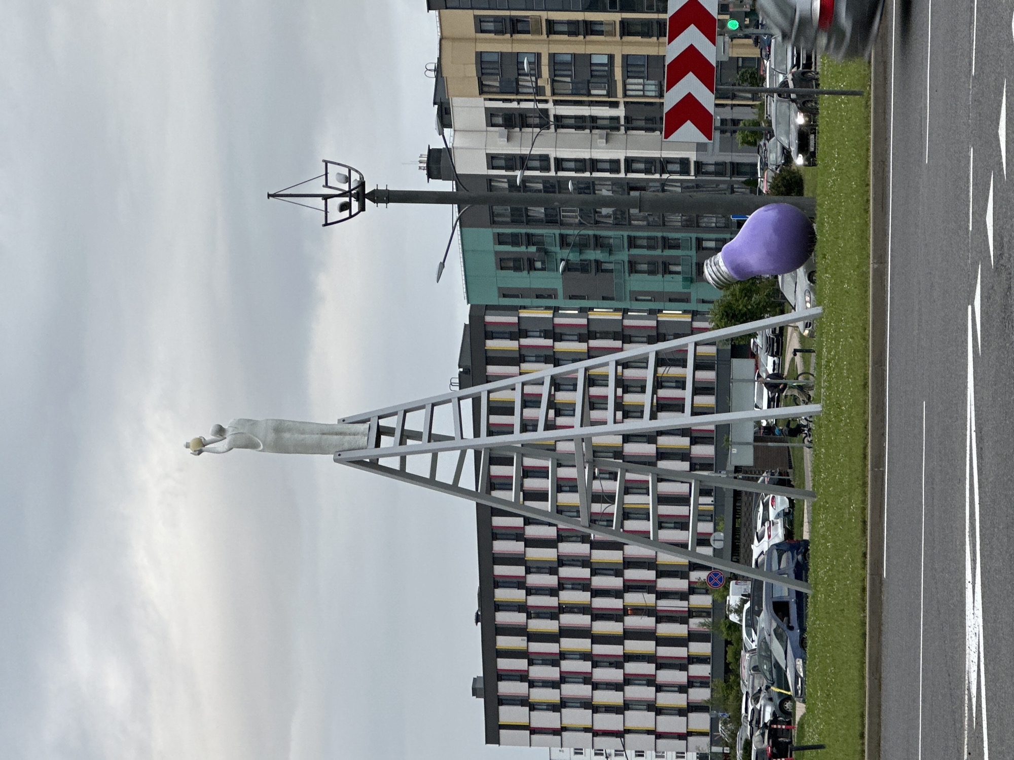 The image depicts an outdoor scene with a sculpture of a person reaching upwards. This sculpture is positioned on a large, abstract structure that resembles a ladder or an A-frame. The sculpture is made of a light-colored material, possibly stone or concrete, and appears to be holding a small, spherical object in its raised hand.
In the background, there are modern buildings with distinctive checkered facades in shades of white, yellow, and red. A large, vibrant purple lightbulb sculpture is visible on the grassy area beside a road. The road has white lane markings and several parked cars. The sky is overcast and grey, suggesting a cloudy day. The overall mood is somewhat somber due to the weather but brightened by the colorful sculptures and buildings.
The scene appears to be in an urban or semi-urban environment, possibly a park or public space adjacent to residential or commercial buildings and a road. The time of day is not clearly discernible due to the overcast sky, but it seems to be daytime. There are no discernible text elements in the image, other than a round blue and red sign on a pole near the parked cars.