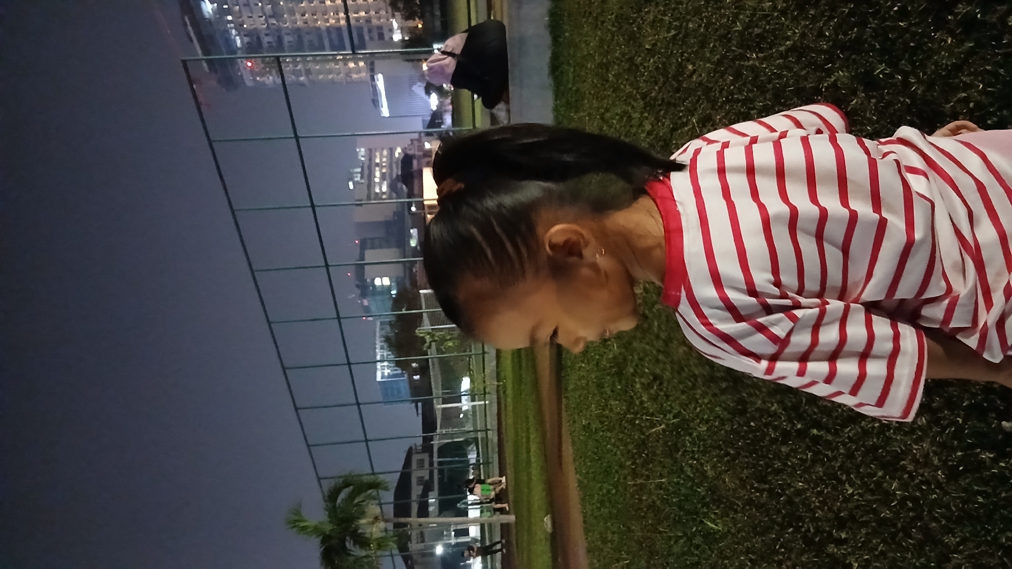 A young girl with dark hair tied in a ponytail, wearing a white t-shirt with red horizontal stripes, is seen from behind, facing left. She is standing on a grassy area in what appears to be an outdoor sports field or park at night. In the background, a fence separates the field from a cityscape with illuminated buildings and a tall construction crane. There are a few other indistinct figures in the mid-ground, and a palm tree to the left. The lighting suggests an urban environment in Medan, Indonesia.